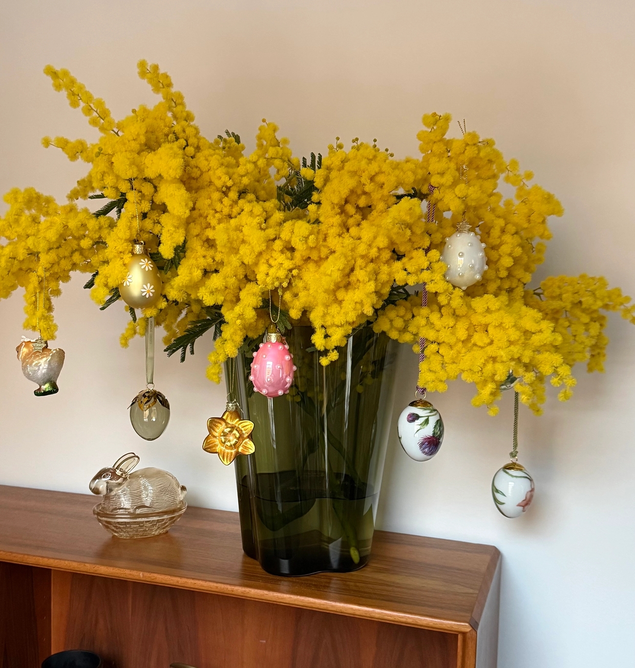 Bright yellow mimosa flowers in a green vase, decorated with Easter ornaments, sit on a wooden shelf with a glass bunny figurine.