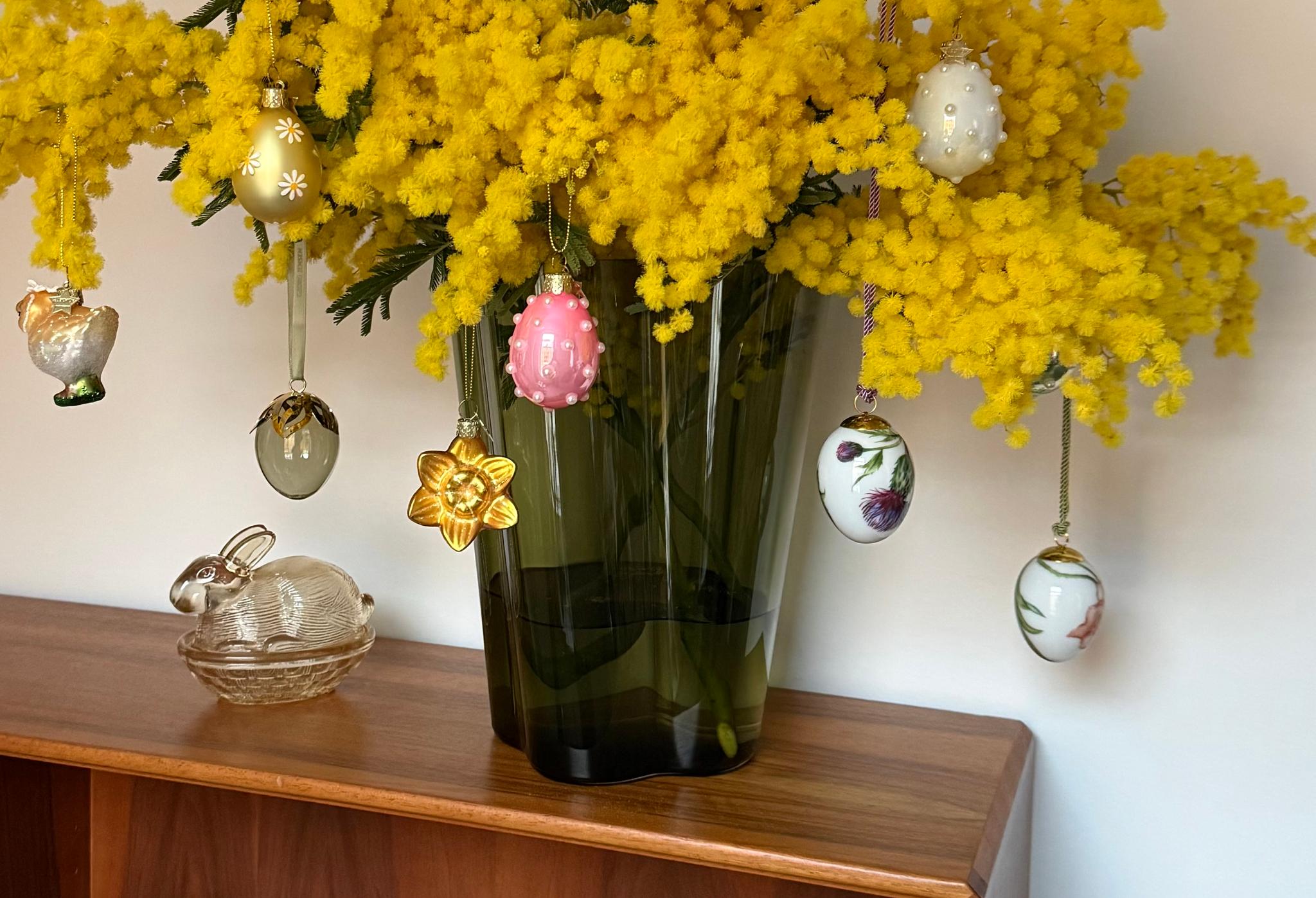 Keltaisia mimosakukkia maljakossa, jota koristavat pääsiäiskoristeet, lasikanin vieressä puupinnalla.