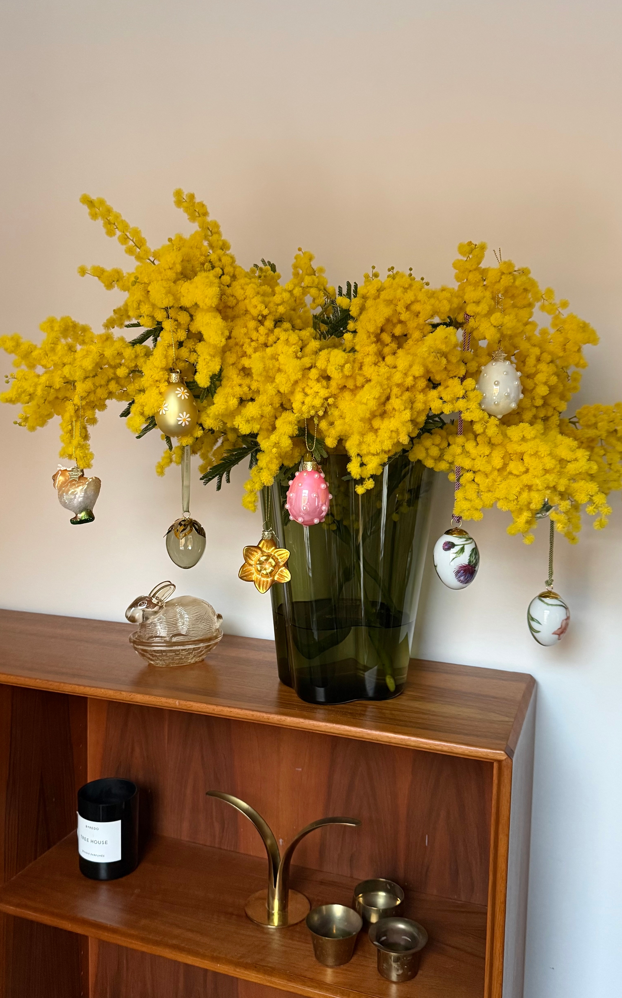 En bukett med knallgule mimosablomster i en vase, pyntet med hengende påskeegg