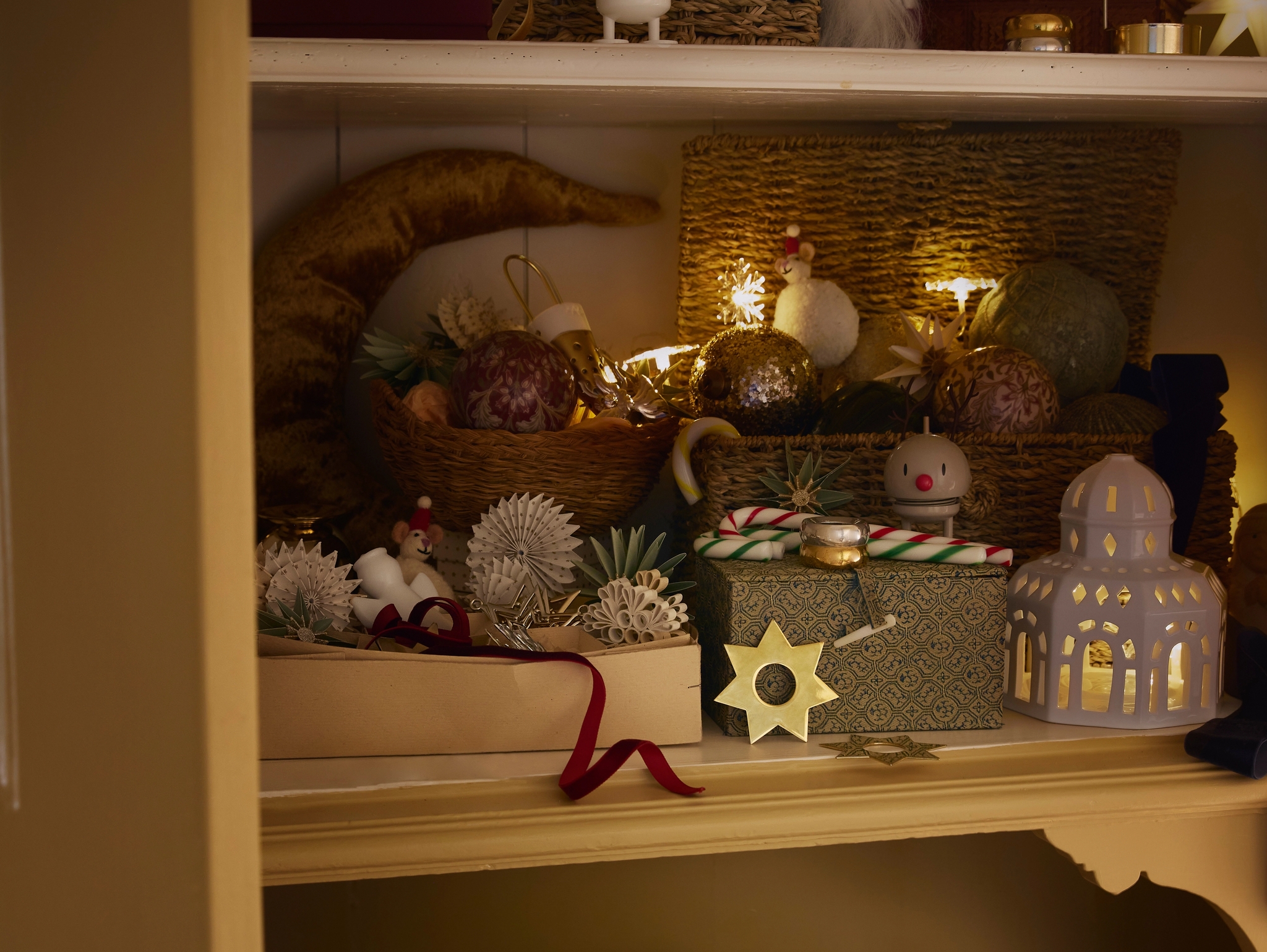 Vitriinikaappi täynnä jouluisia koristeita – hehkuvia tähtivaloja, kuusenkoristeita, lahjoja ja pieniä figuureja – luoden lämpimän ja kodikkaan tunnelman.