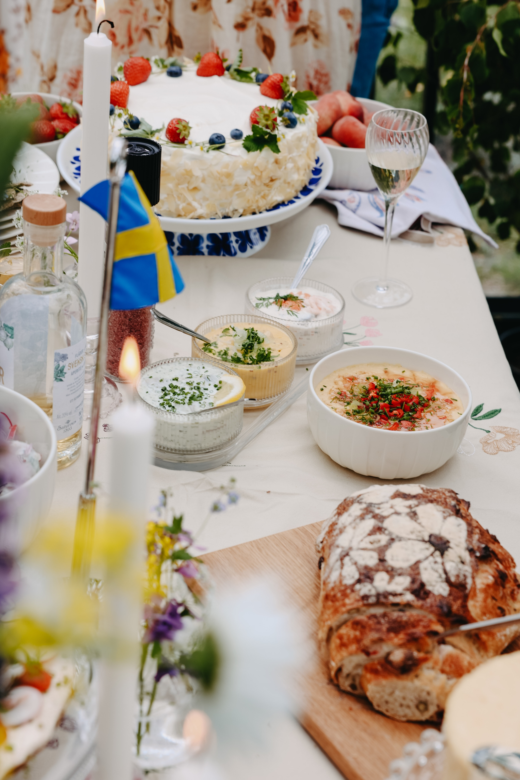 Somrig svensk festdukning med jordgubbstårta, bröd, sillröror och liten svensk flagga på bordet.