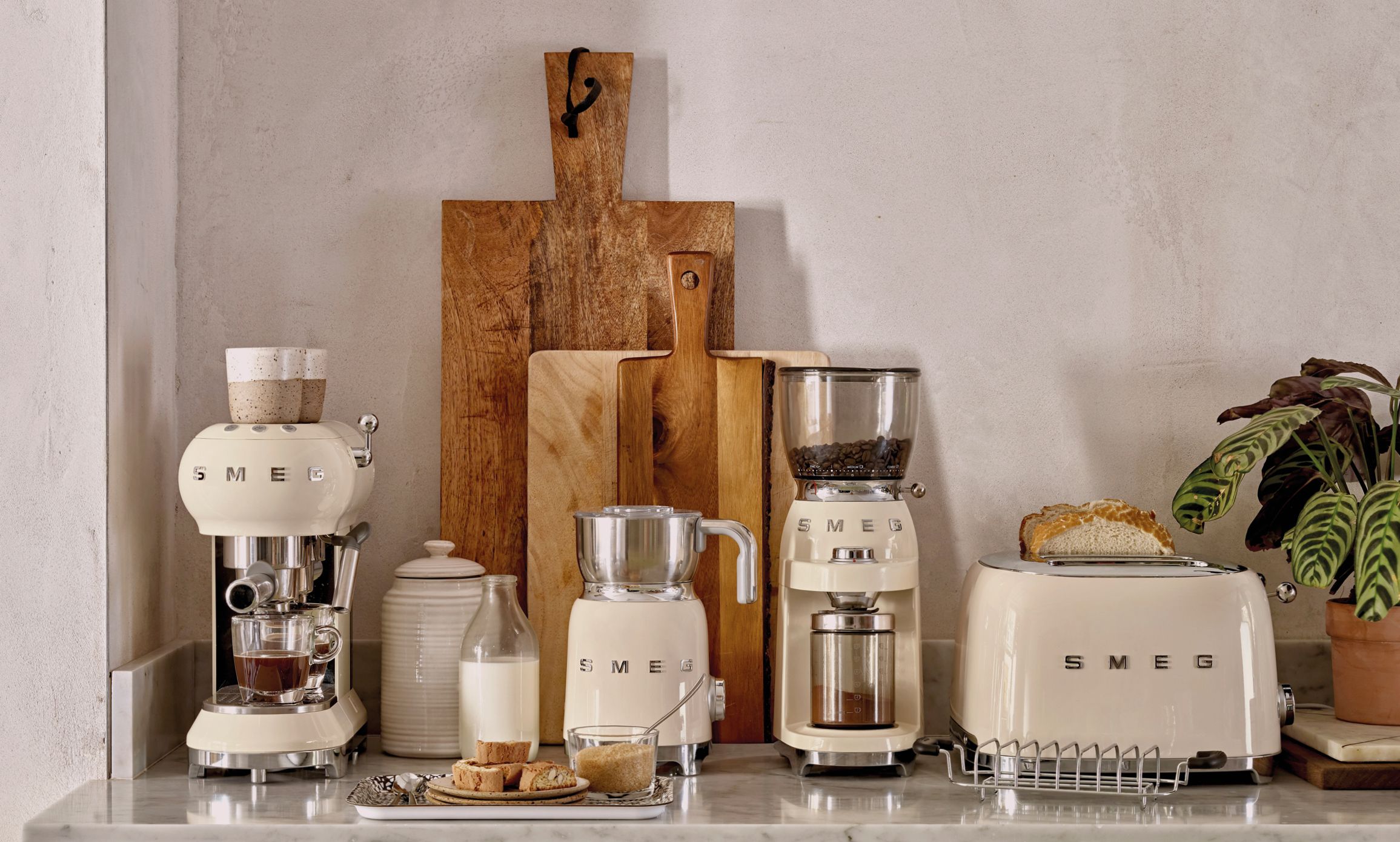 Stilren köksbänk med krämfärgade SMEG-köksmaskiner, brödrost, kaffemaskin och träskärbrädor mot en ljus vägg.