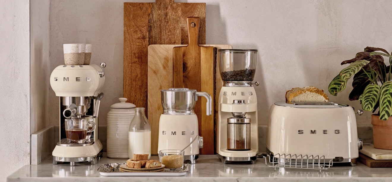 Stilren köksbänk med krämfärgade SMEG-köksmaskiner, brödrost, kaffemaskin och skärbrädor i trä mot en ljus vägg.