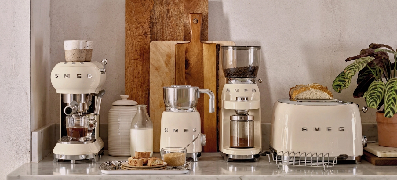 Stilren köksbänk med krämfärgade SMEG-köksmaskiner, brödrost, kaffemaskin och träskärbrädor mot en ljus vägg.
