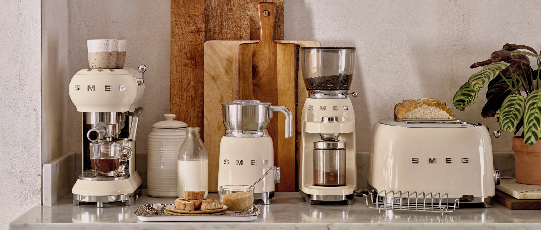 Stilren kjøkkenbenk med kremfargede SMEG-apparater, kaffemaskin, brødrister og kaffekvern foran trefjøler og en grønn potteplante.