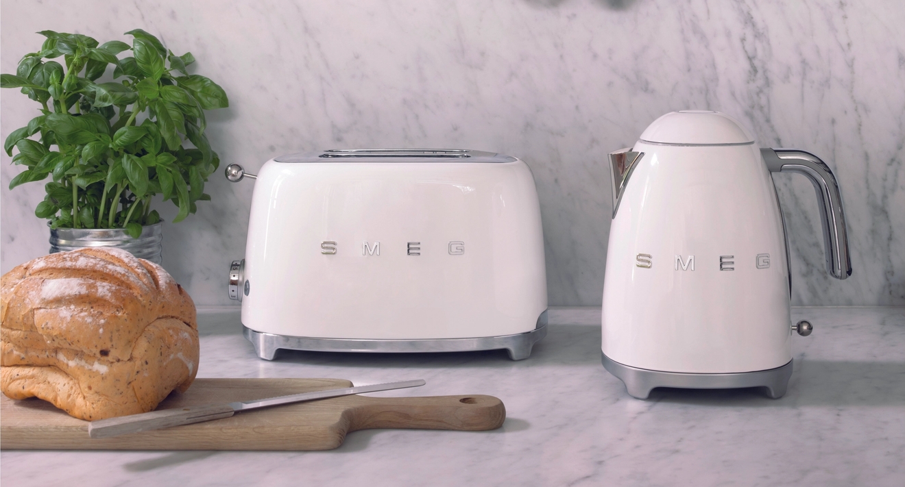 Stilren köksbänk i marmor med vita SMEG-brödrost och vattenkokare, färskt bröd och basilika framför mörk panelvägg med kopparkastrull och träredskap.