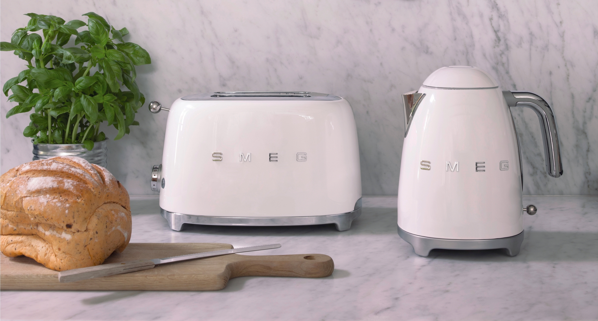 Valkoinen Smeg-leivänpaahdin ja vedenkeitin marmoritasolla, jossa on tuoretta basilikaa, leipää ja yläpuolella roikkuvia kuparisia keittiövälineitä.