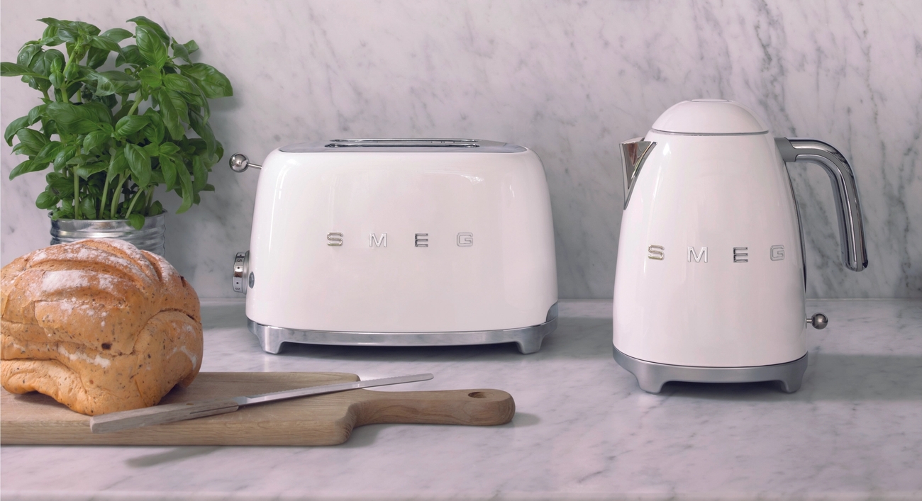 Hvit Smeg-brødrister og vannkoker på en kjøkkenbenk i marmor med fersk basilikum, brød og kjøkkenutstyr i kobber hengende over.