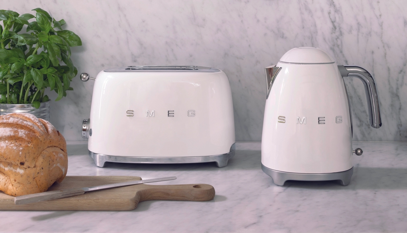 Vit Smeg-brödrost och vattenkokare på en marmorköksbänk med färsk basilika, bröd och köksredskap i koppar som hänger ovanför.