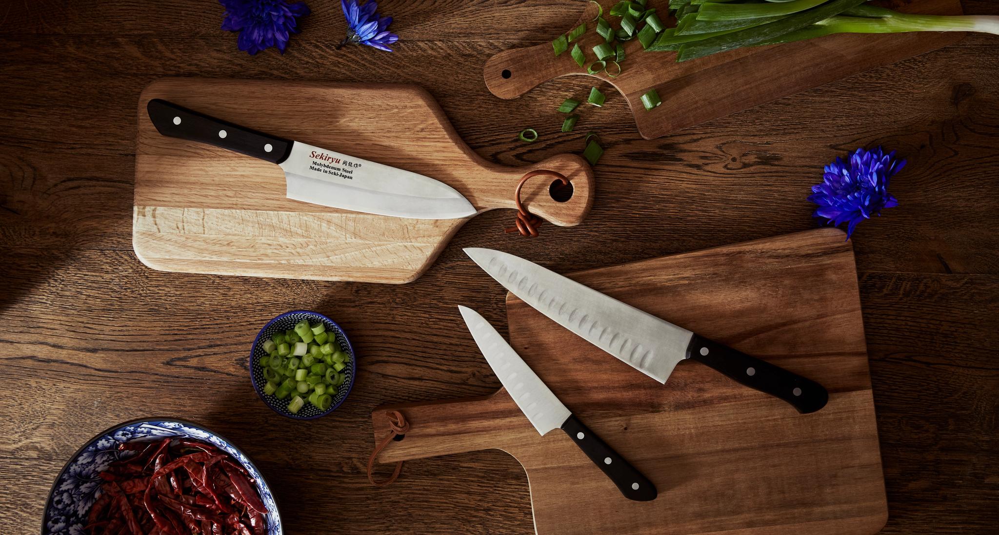 Översiktsvy uppifrån av tre skärbrädor i trä med köksknivar, salladslök, torkade röda chilifrukter och blå blommor på en mörk träyta.