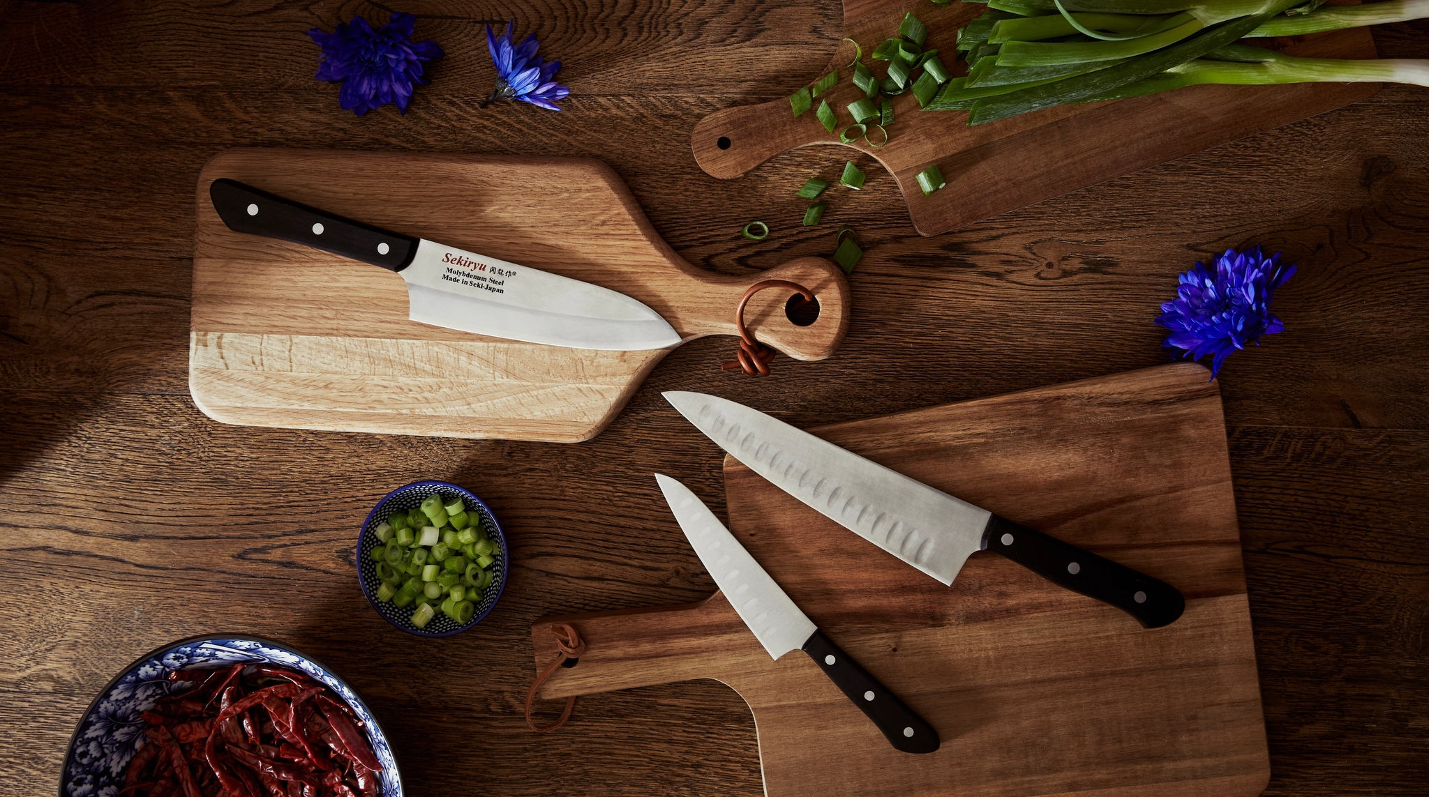 Översiktsvy uppifrån av tre skärbrädor i trä med köksknivar, salladslök, torkade röda chilifrukter och blå blommor på en mörk träyta