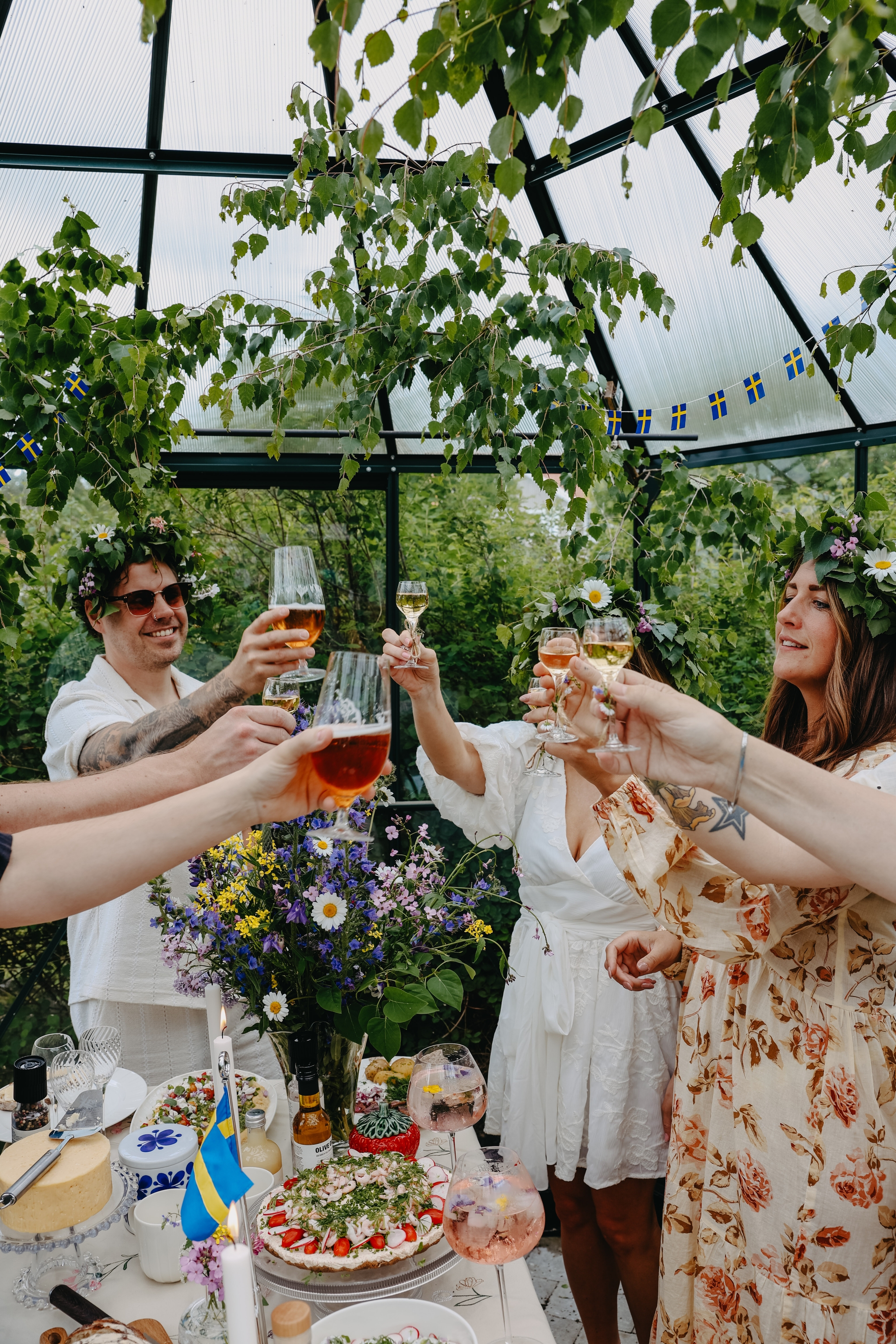 Vänner med blomsterkransar skålar runt ett midsommarbord fullt av svensk sommar mat och dryck i ett grönt växthus dekorerat med björkris och svenska flaggor.