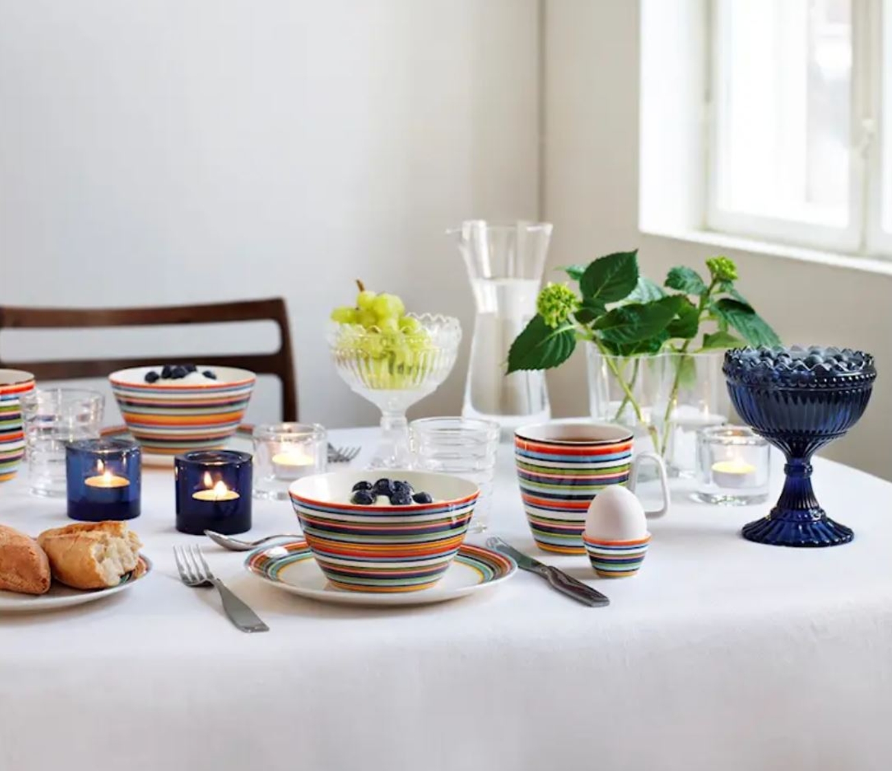 A breakfast table set with colorful striped dishes, blueberries, grapes, bread, an egg, and lit candles.