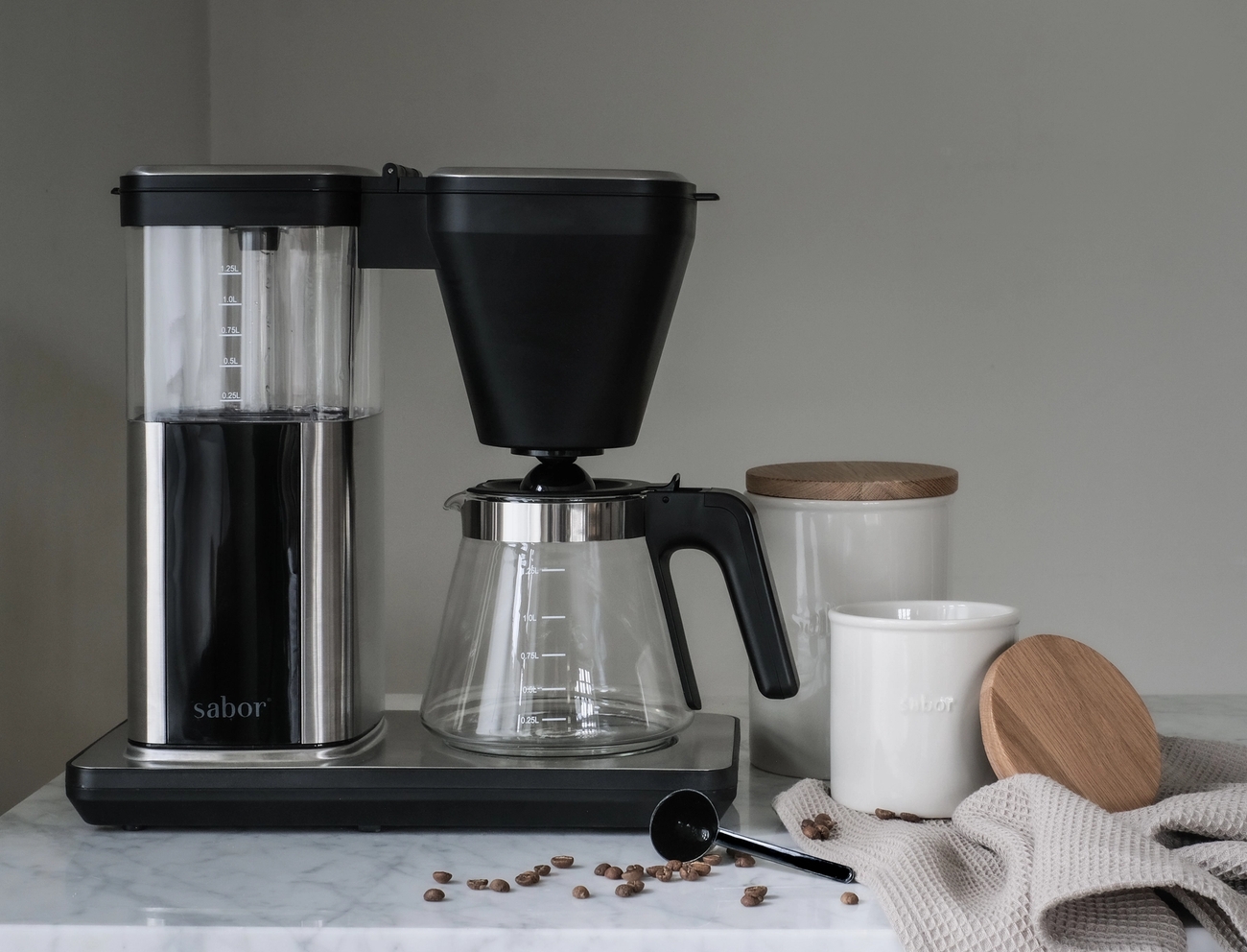 A Sabor coffee maker, coffee beans, and white ceramic canisters with wooden lids on a marble countertop.