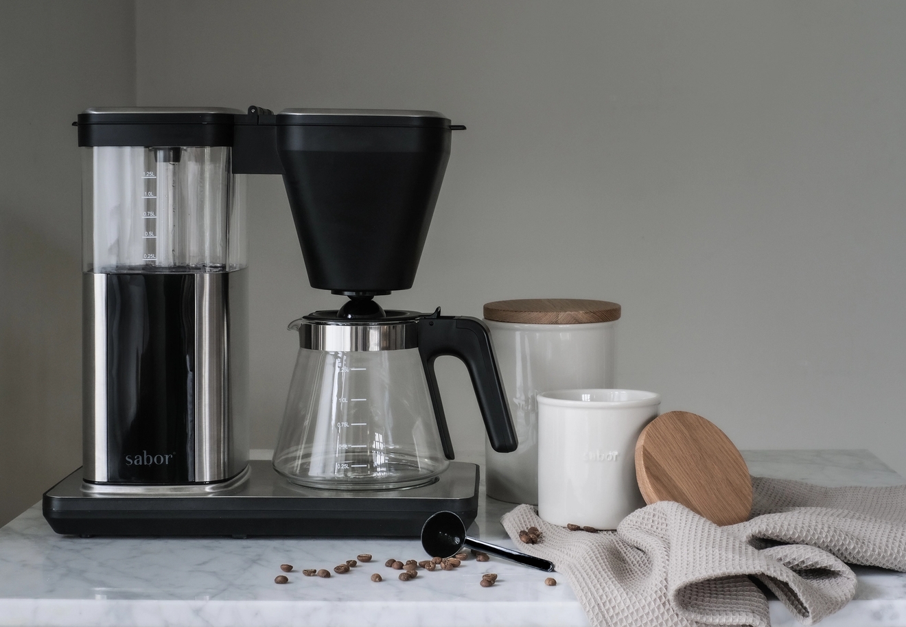 En Sabor-kaffebryggare och karaff på en marmorbänkskiva med kaffebönor, en sked, två vita behållare och en beige handduk.