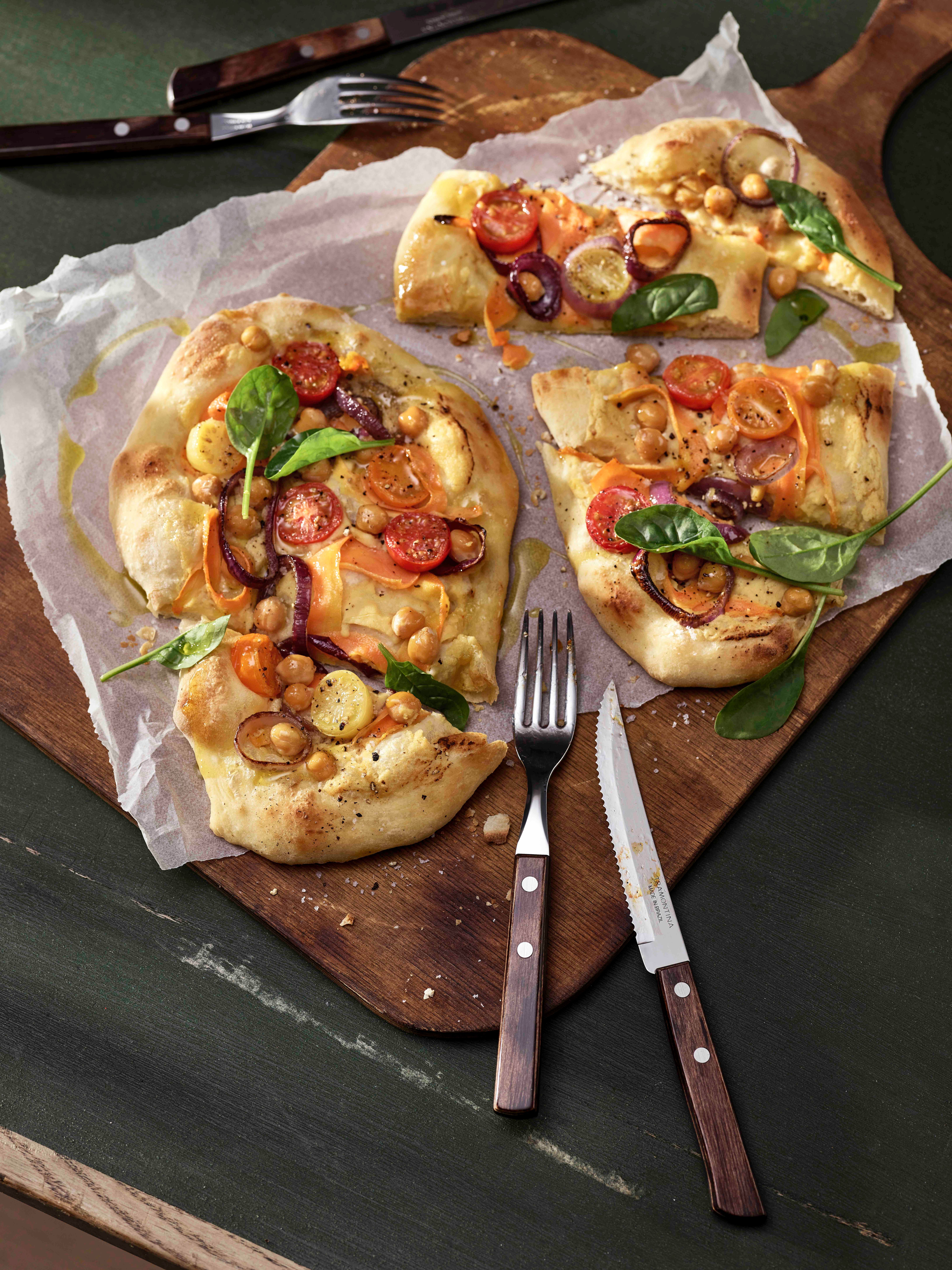 Tre bitar av ett flatbröd med ljus skorpa toppat med körsbärstomater, kikärtor, rödlök och spenat, serverade på en träbräda med gaffel och kniv