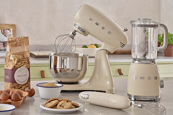 Krämfärgade Smeg köksmaskiner står på en köksbänk omgivna av ingredienser som ägg, mjöl och kakor, redo för bakning.