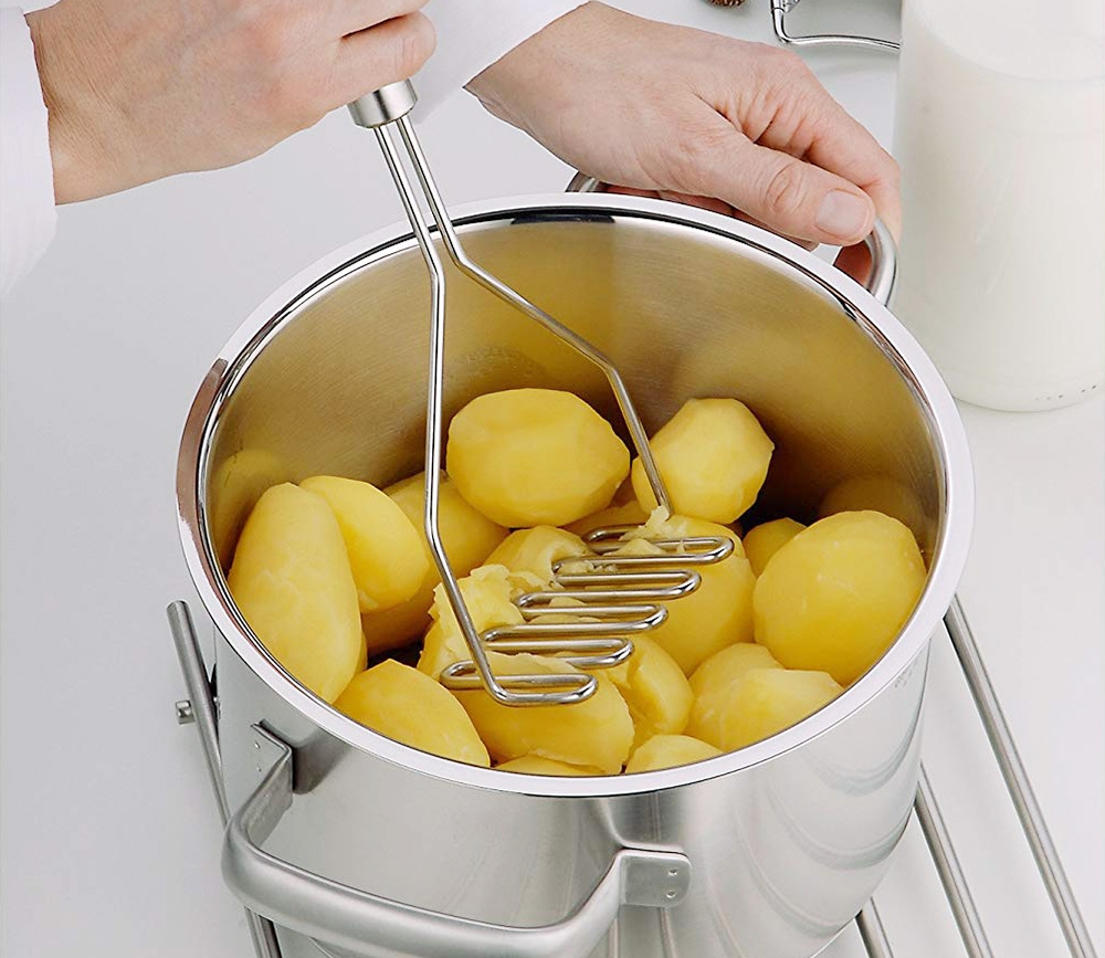 En person använder en potatisstöt för att mosa kokta potatisar i en gryta.