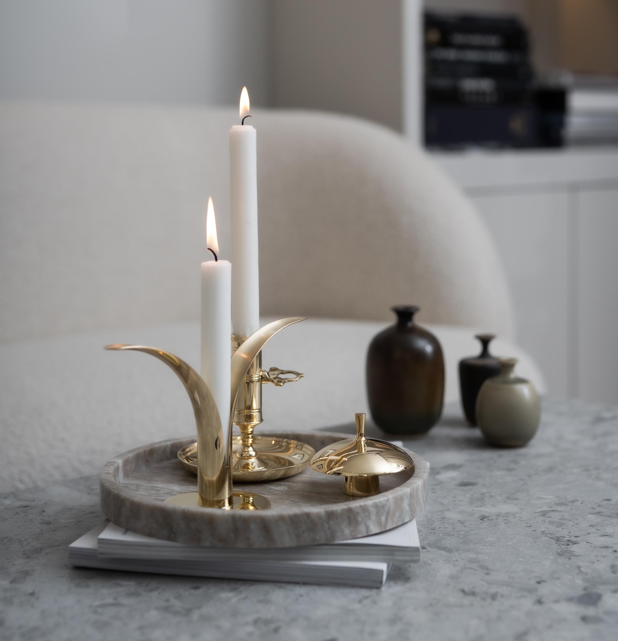 Två tända vita ljus i mässingsljusstakar på ett marmorfat, placerade på ett grått marmorbord med suddig modern inredning i bakgrunden.