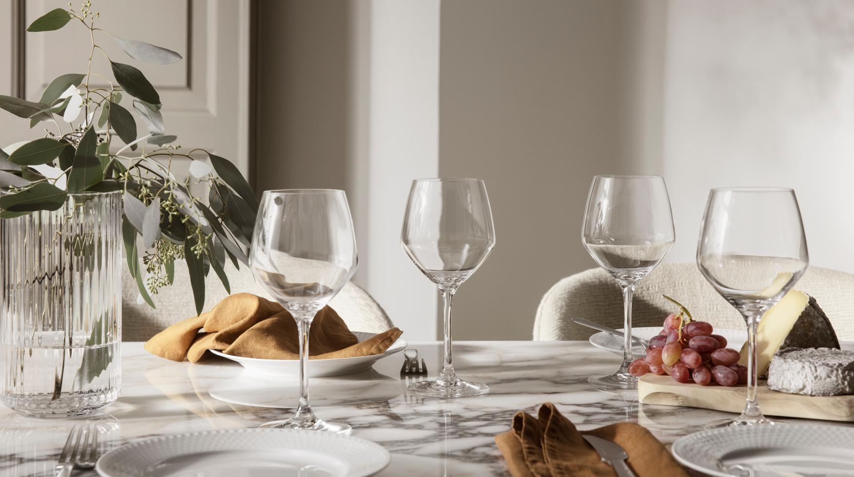 Elegant, lys borddekking på marmorbord med vinglass, hvite tallerkener, linservietter, grønne kvister i vase og ostefat med druer.