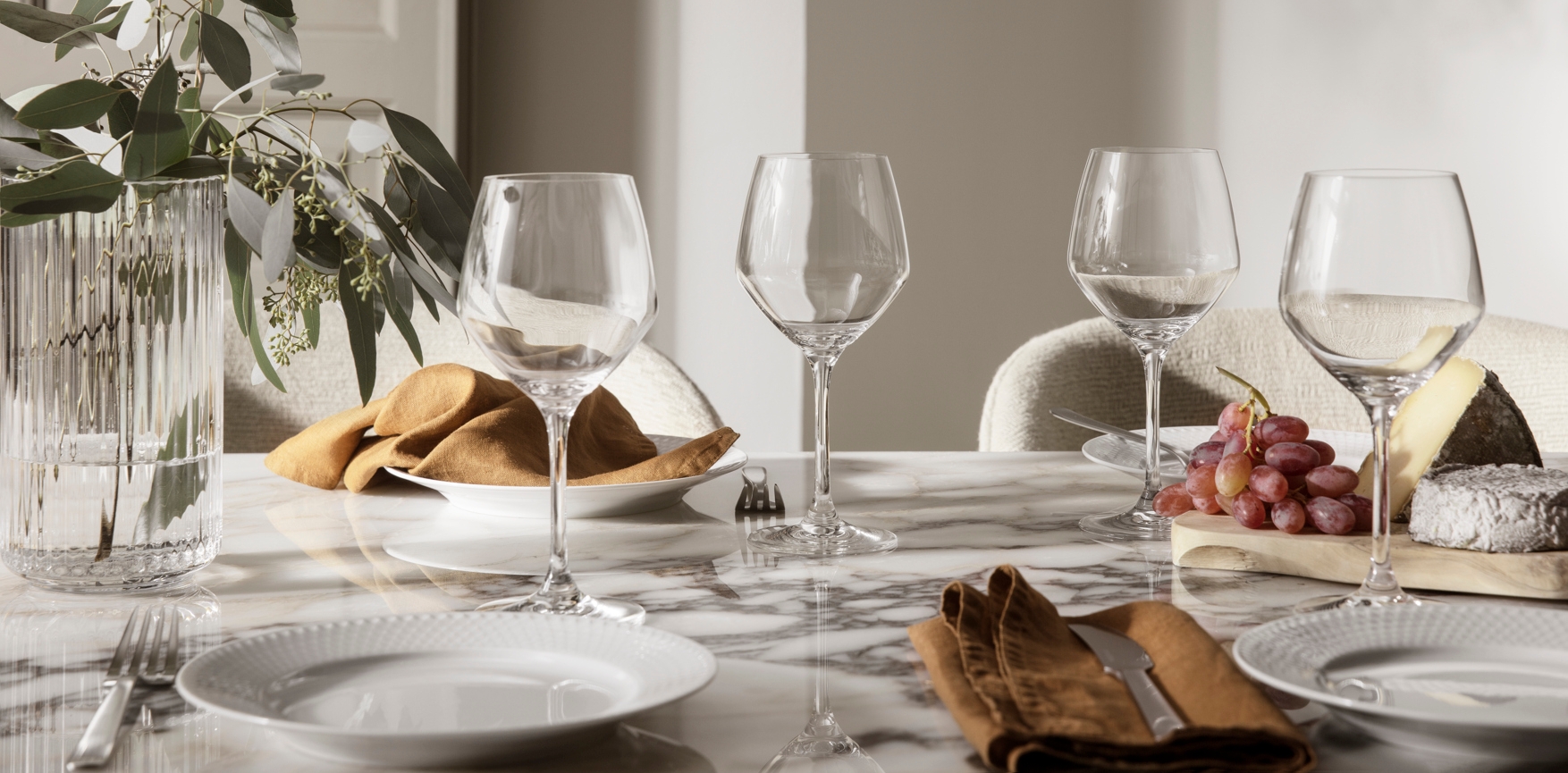 Elegant dukning på marmorbord med vinglas, eukalyptus, bröd, vindruvor och ostar.