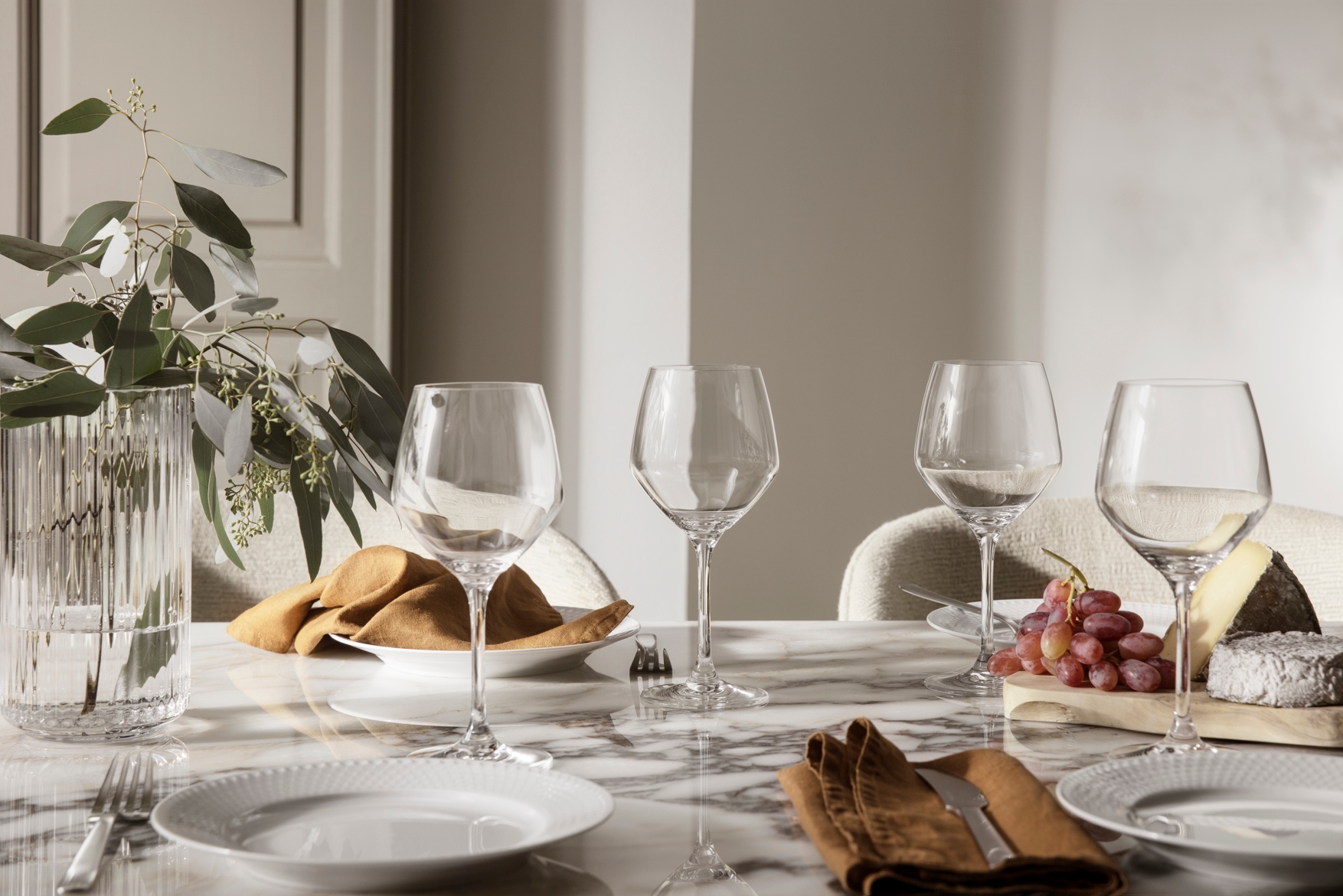Elegant dekket marmorbord med vinglass, hvite tallerkener, linservietter, ost og druer i et lyst, moderne spiserom.