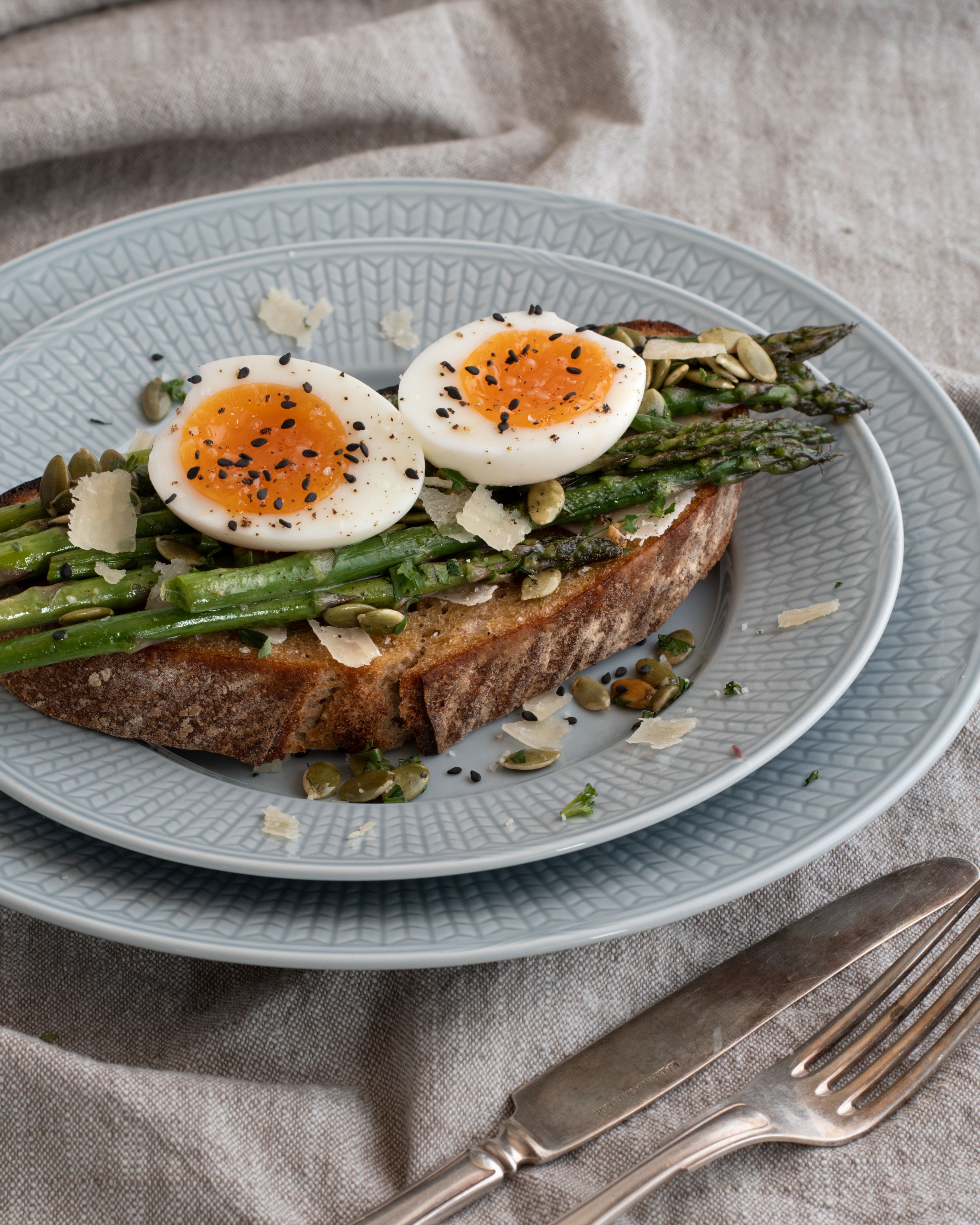 Rostat bröd toppat med ugnsrostad sparris, två halverade löskokta ägg strödda med svarta sesamfrön, pumpafrön och riven ost, serverat på en blå tallrik.