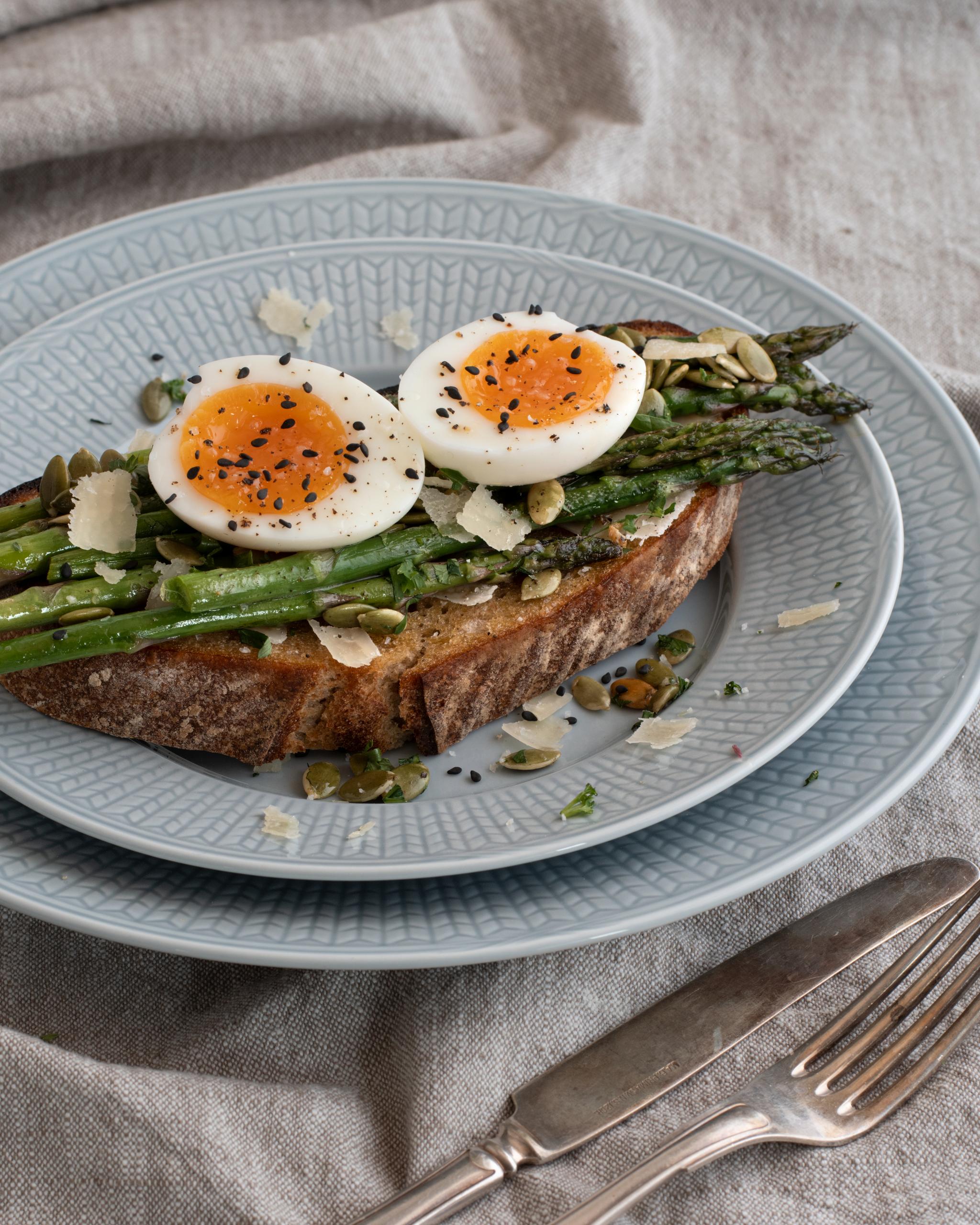 Sparris- och löskokt äggtoast, garnerad med frön och ost, på en ljusblå tallrik.