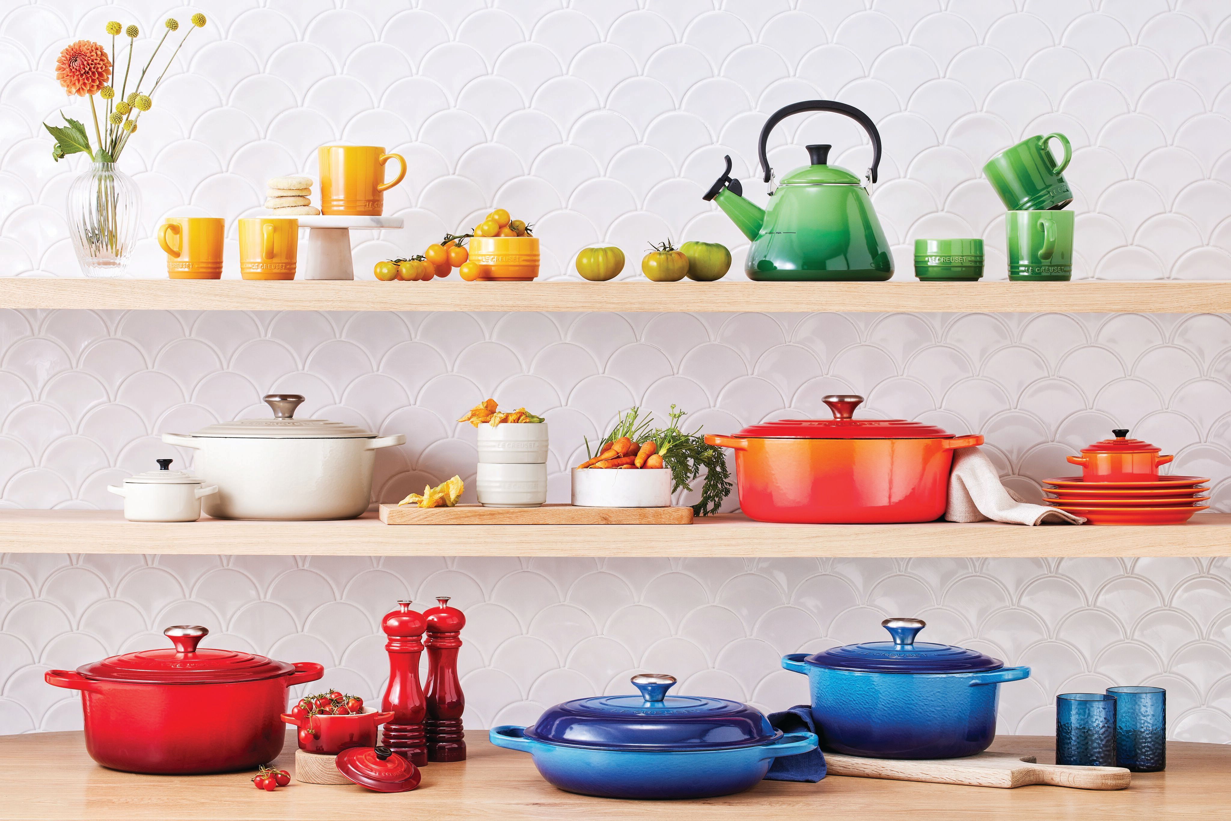 Färgglada Le Creuset köksredskap och tillbehör visas på trähyllor mot en vit, vågig kakelplatta.