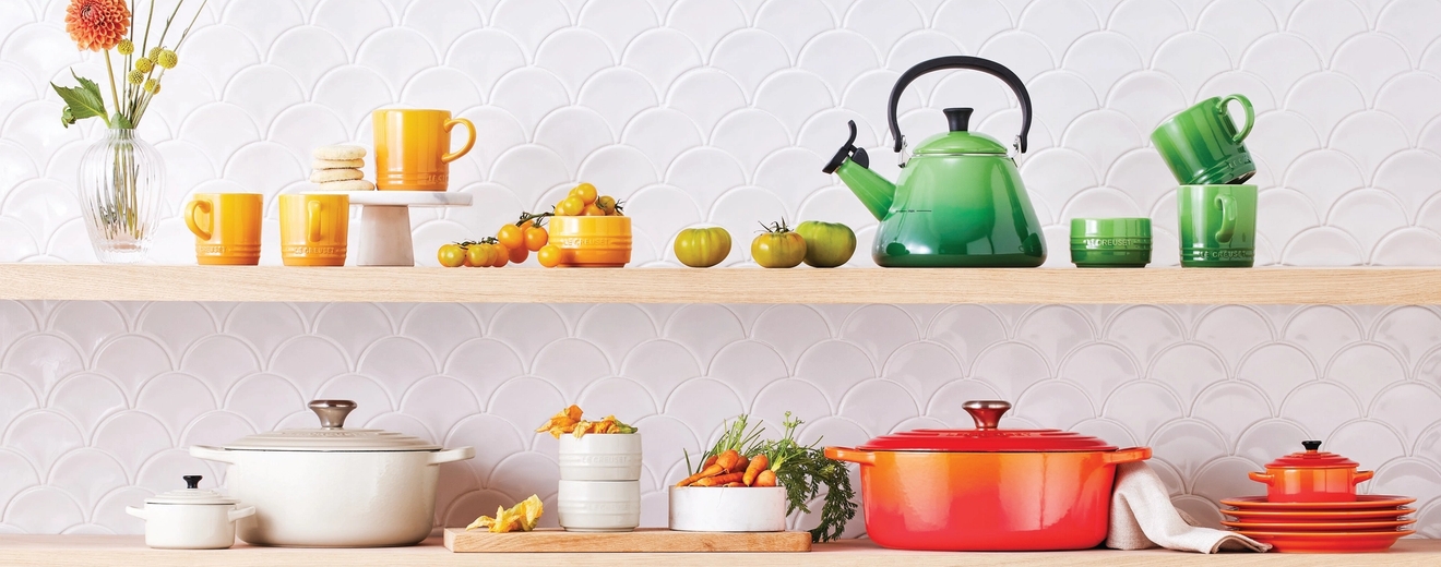 Färgglada Le Creuset köksredskap och tillbehör visas på trähyllor mot en vit, vågig kakelplatta.