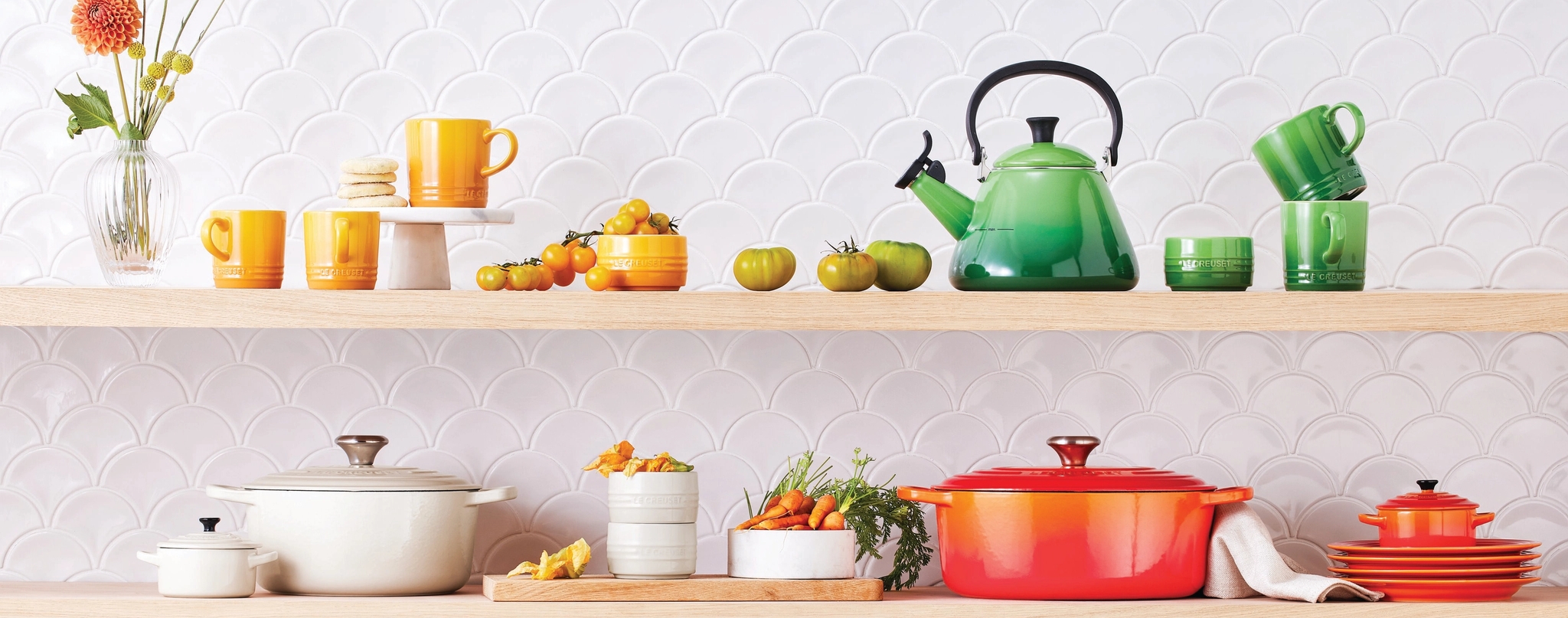 Färgglada Le Creuset köksredskap och tillbehör visas på trähyllor mot en vit, vågig kakelplatta.