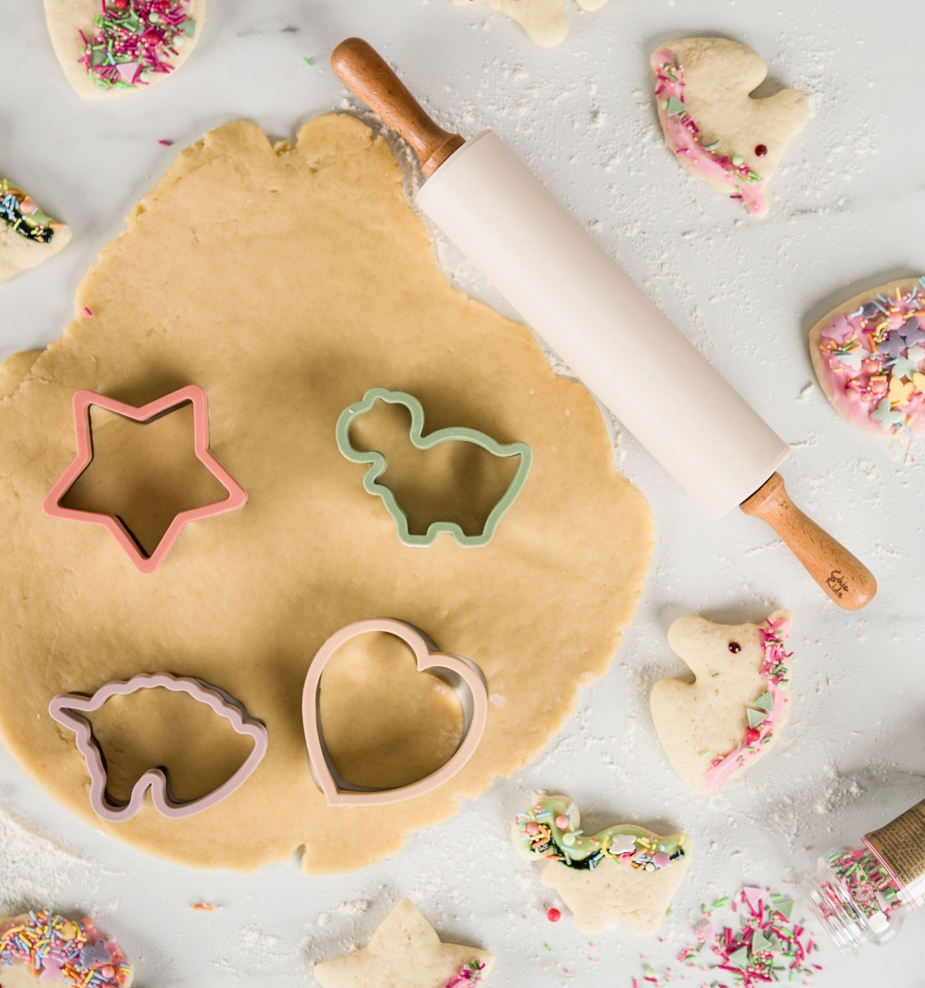Chic Kids Utrullad kakdeg med färgglada pepparkaksformar, kavel och dekorerade småkakor på ett mjölat bakbord.