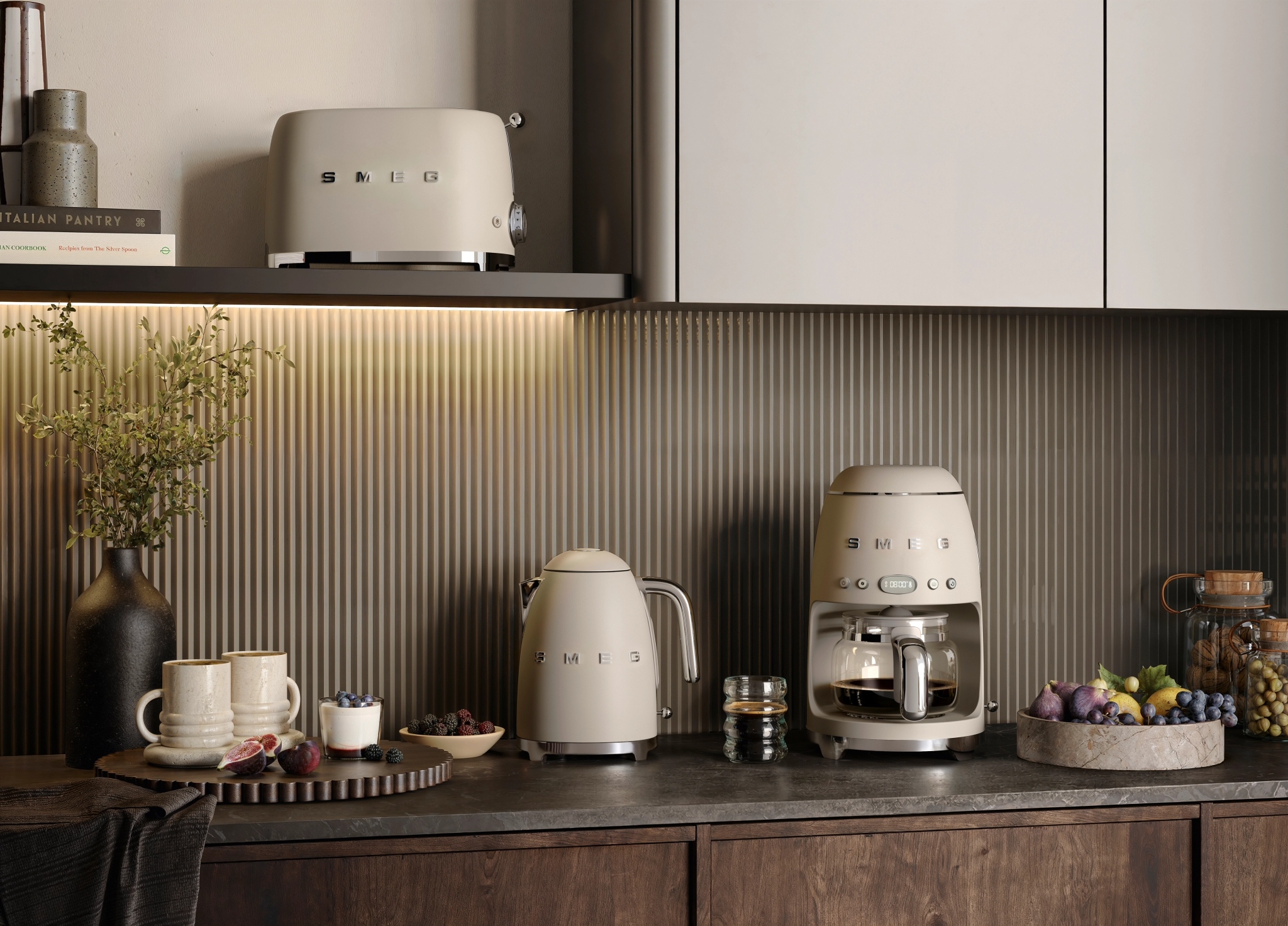Stilrent kök med beige Smeg-kaffebryggare, vattenkokare och brödrost bland frukt och dekorativa detaljer.