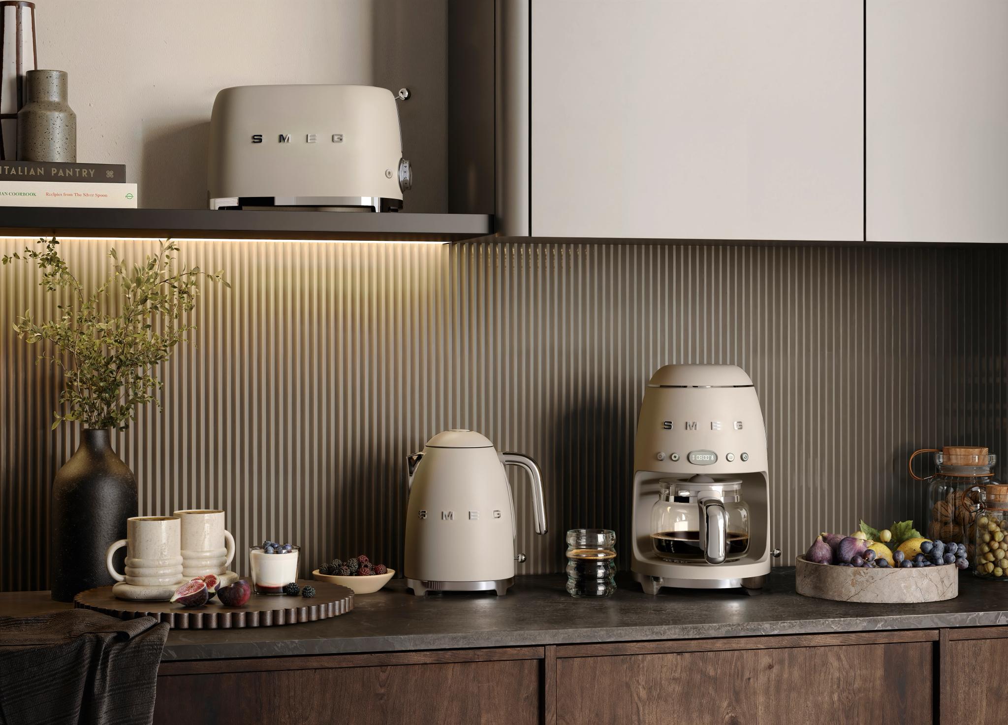 Stilrent kjøkken med beige Smeg-kaffetrakter, vannkoker og brødrister omgitt av frukt og keramikk.