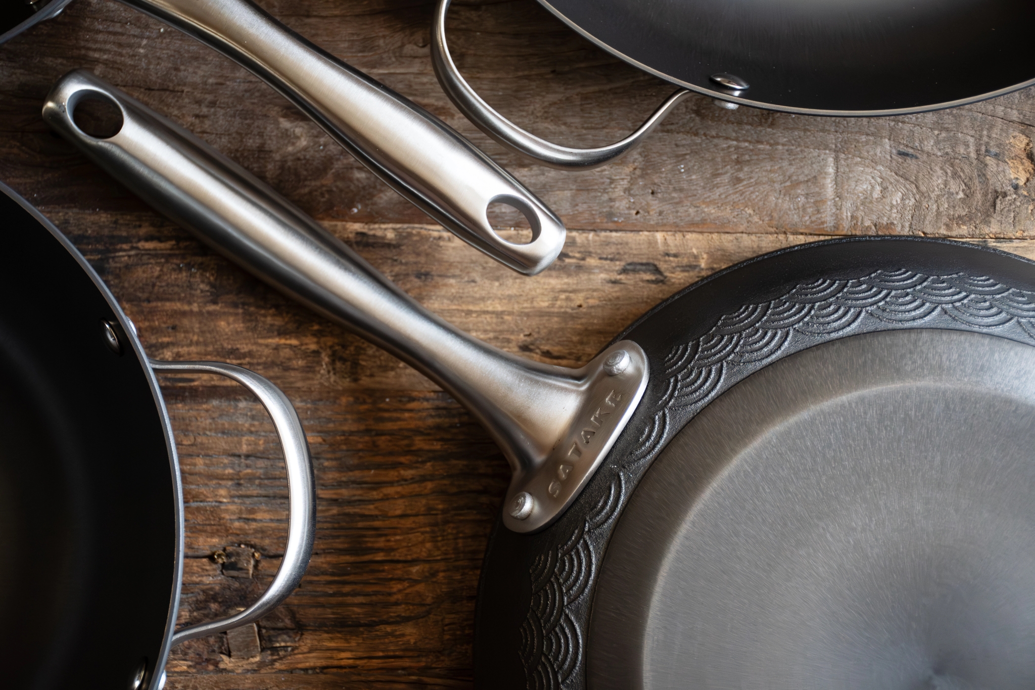 Ovanifrånvy av flera mörka kokpannor och grytor med silverhandtag på trä, en pannas undersida visar ett dekorativt vågmönster.