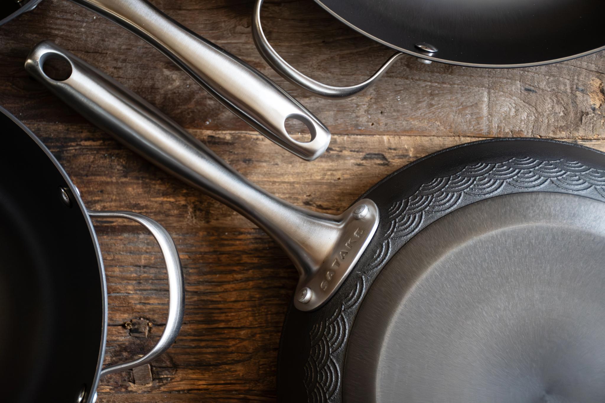 Ovanifrånvy av flera mörka kokpannor och grytor med silverhandtag på trä, en pannas undersida visar ett dekorativt vågmönster.