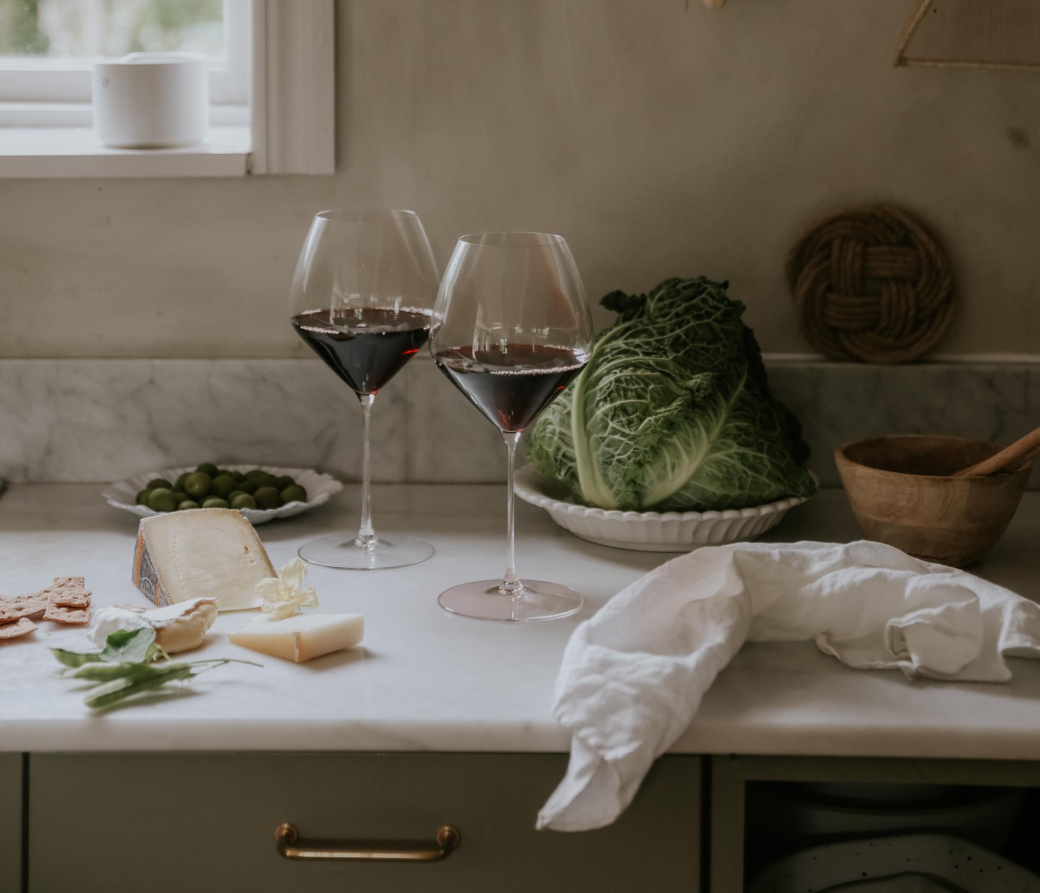 Två glas rött vin står på en köksbänk med savojkål, ost och oliver i ett rustikt kök.