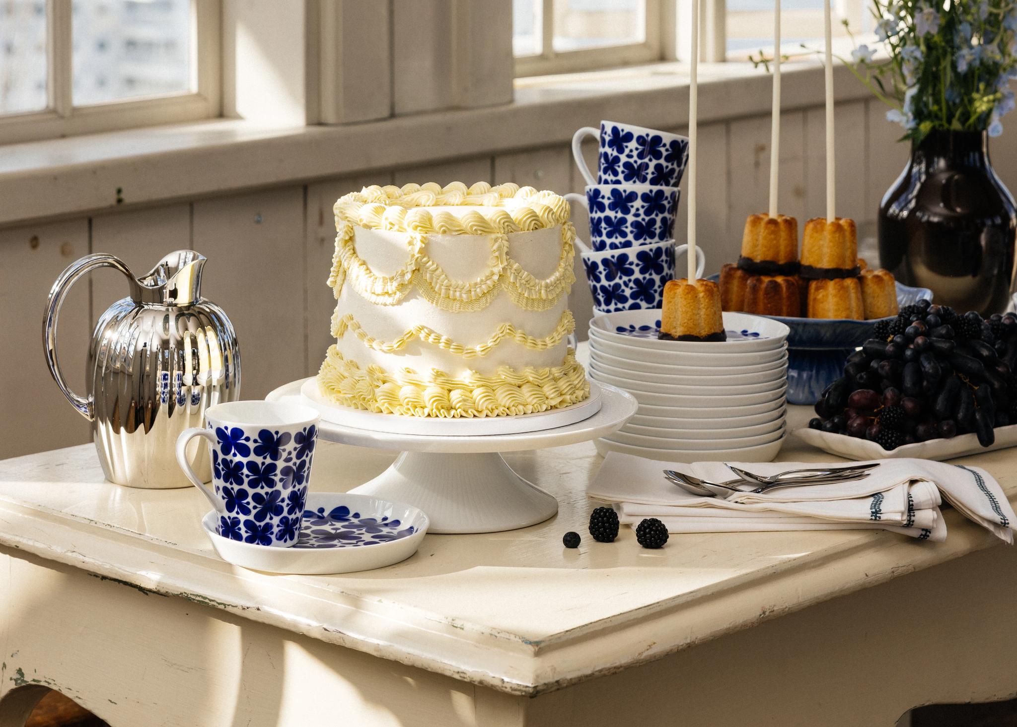 Ett dessertbord med en vit tårta, små gyllene kakor på pinnar, blå blommönstrade muggar, en silverkanna och vindruvor