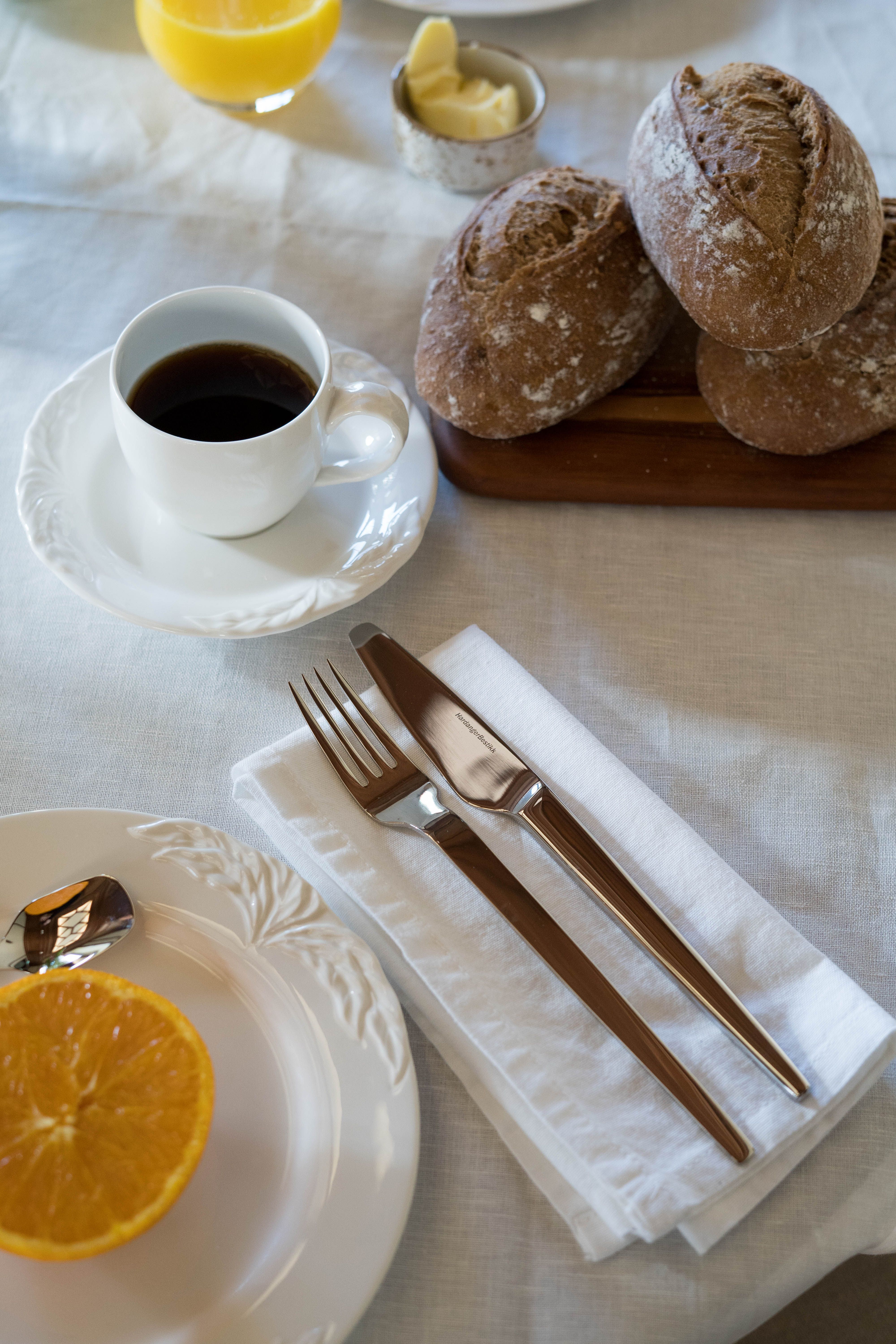 Aamiaispöytä katettu kahvilla, maalaismaisilla sämpylöillä, appelsiinimehulla, puolikkaalla appelsiinilla ja aterimilla valkoisella pöytäliinalla.