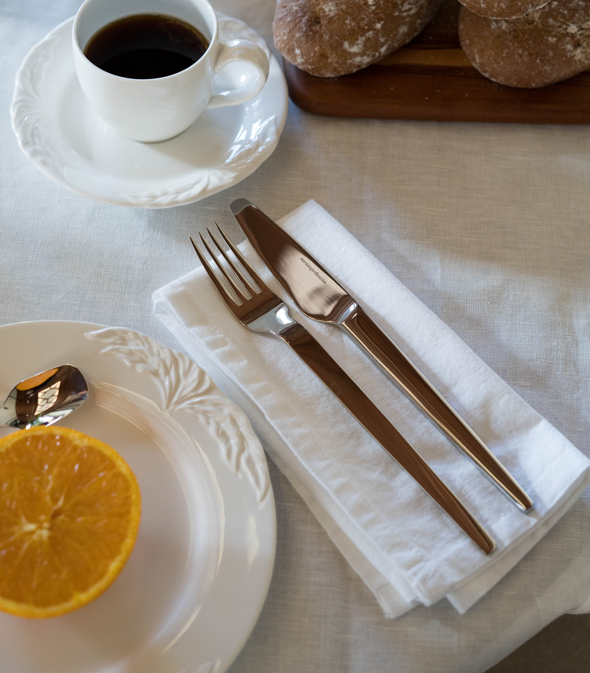 Aamiaispöytä katettu kahvilla, maalaismaisilla sämpylöillä, appelsiinimehulla, puolikkaalla appelsiinilla ja aterimilla valkoisella pöytäliinalla.