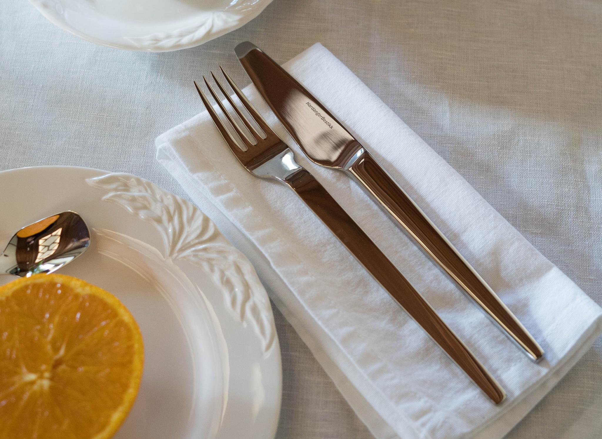 Aamiaispöytä katettu kahvilla, maalaismaisilla sämpylöillä, appelsiinimehulla, puolikkaalla appelsiinilla ja aterimilla valkoisella pöytäliinalla.