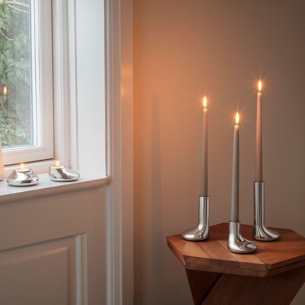 Sytytettyjä kynttilöitä hopeanmuotoisissa kynttilänjaloissa: kaksi tuikkukynttilää ikkunalaudalla ja kolme korkeaa kartiokynttilää saappaanmuotoisissa kynttilänjaloissa puupöydällä.