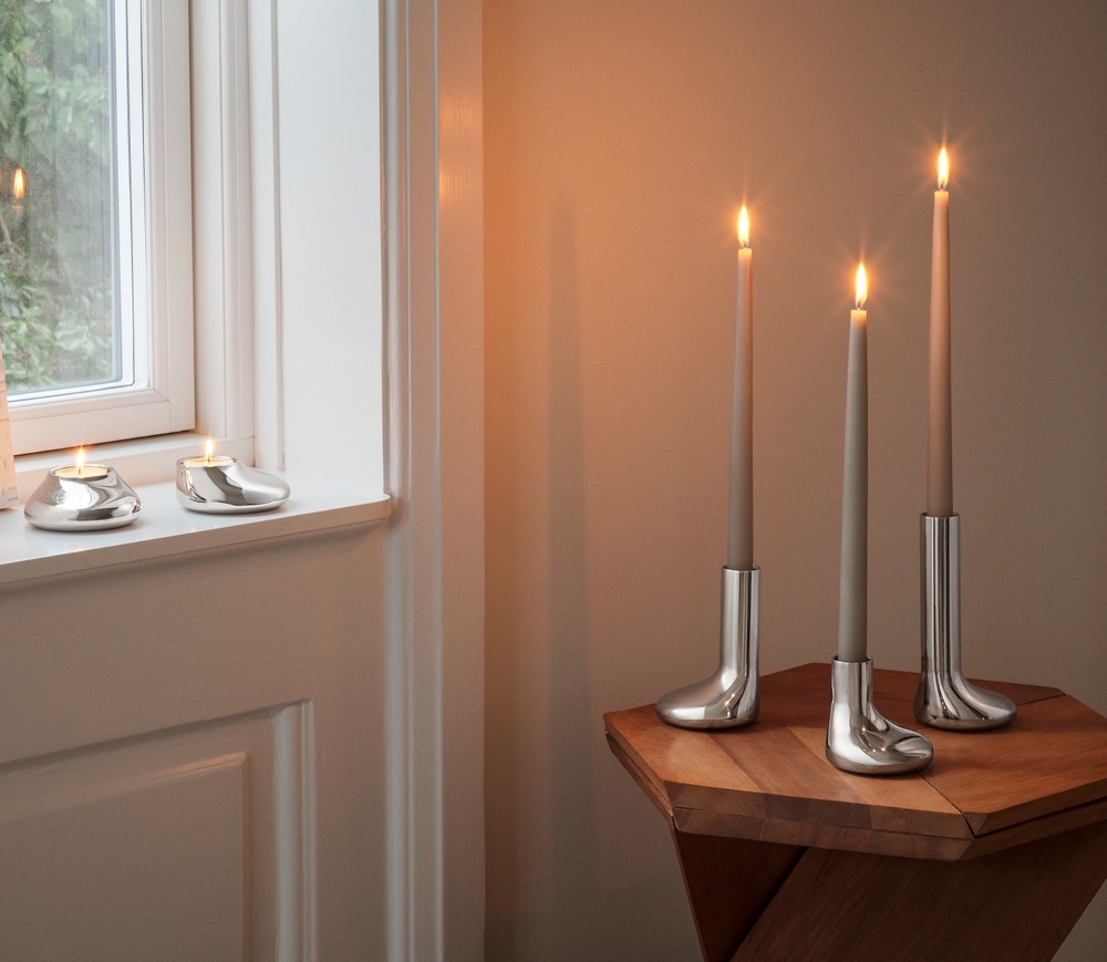 Sytytettyjä kynttilöitä hopeanmuotoisissa kynttilänjaloissa: kaksi tuikkukynttilää ikkunalaudalla ja kolme korkeaa kartiokynttilää saappaanmuotoisissa kynttilänjaloissa puupöydällä.