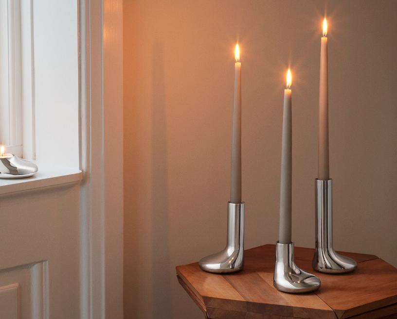 Sytytettyjä kynttilöitä hopeanmuotoisissa kynttilänjaloissa: kaksi tuikkukynttilää ikkunalaudalla ja kolme korkeaa kartiokynttilää saappaanmuotoisissa kynttilänjaloissa puupöydällä.