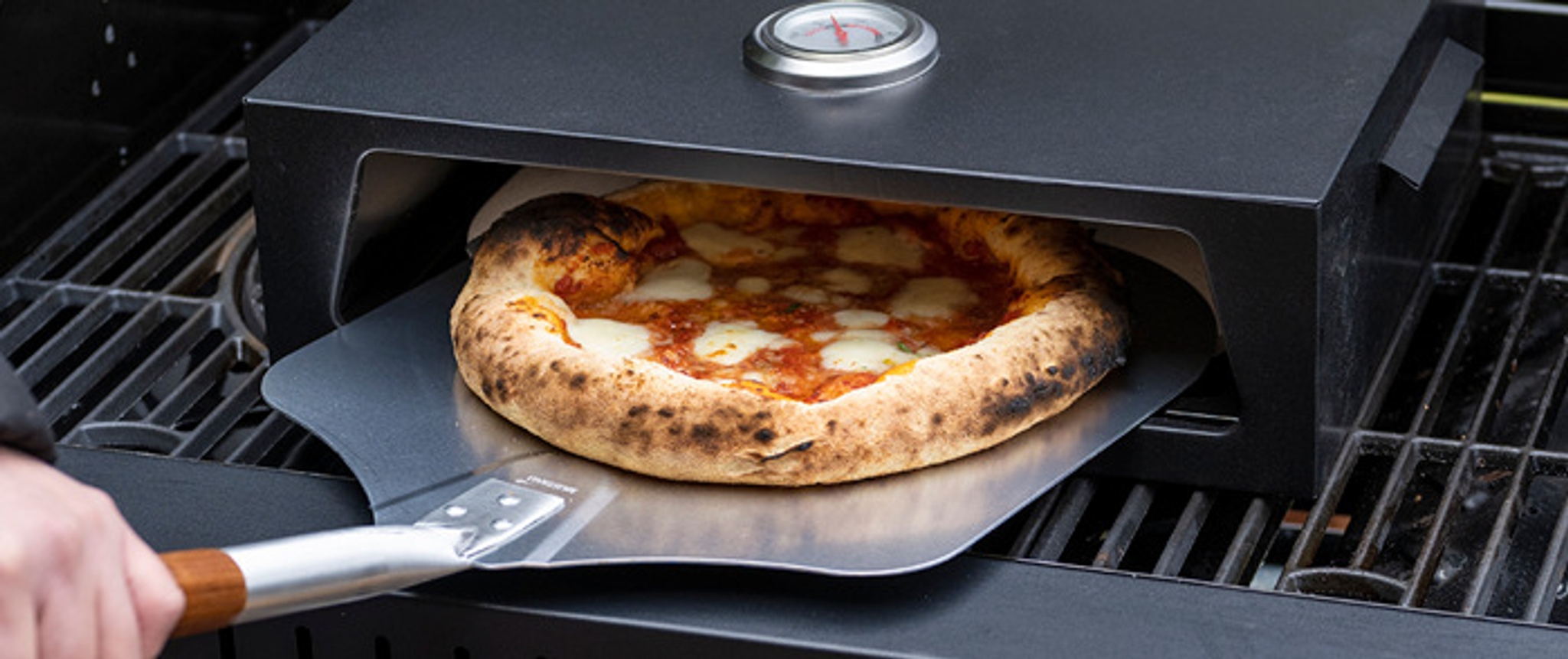 Pizzaugn på grill med nygrillad pizza som tas ut med pizzaspade 