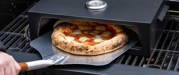 Pizzaugn på grill med nygrillad pizza som tas ut med pizzaspade
