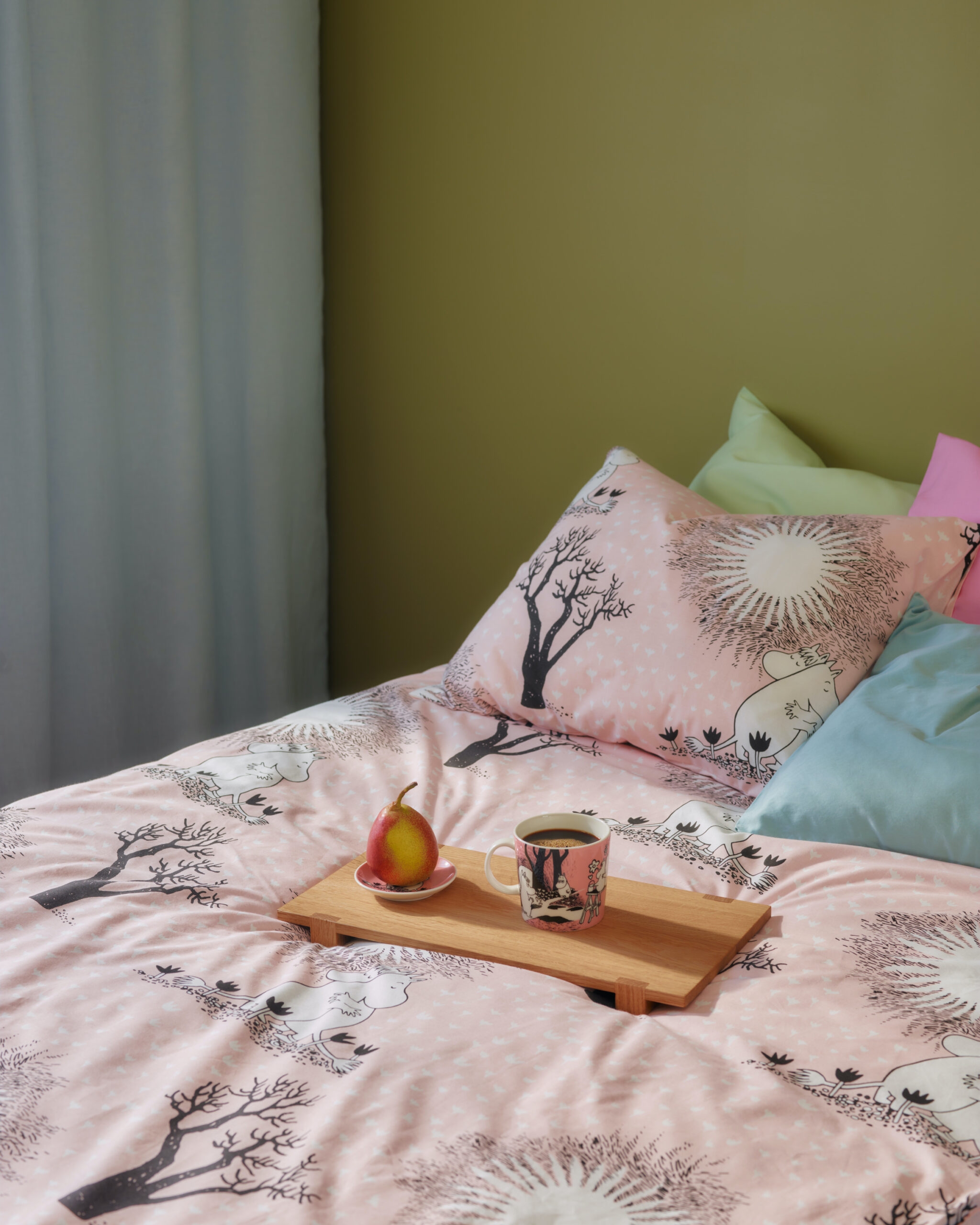 Rosa sengetøy med Mummi-mønster og trær, et trebrett med kaffe og en pære samt fargerike puter på en seng.