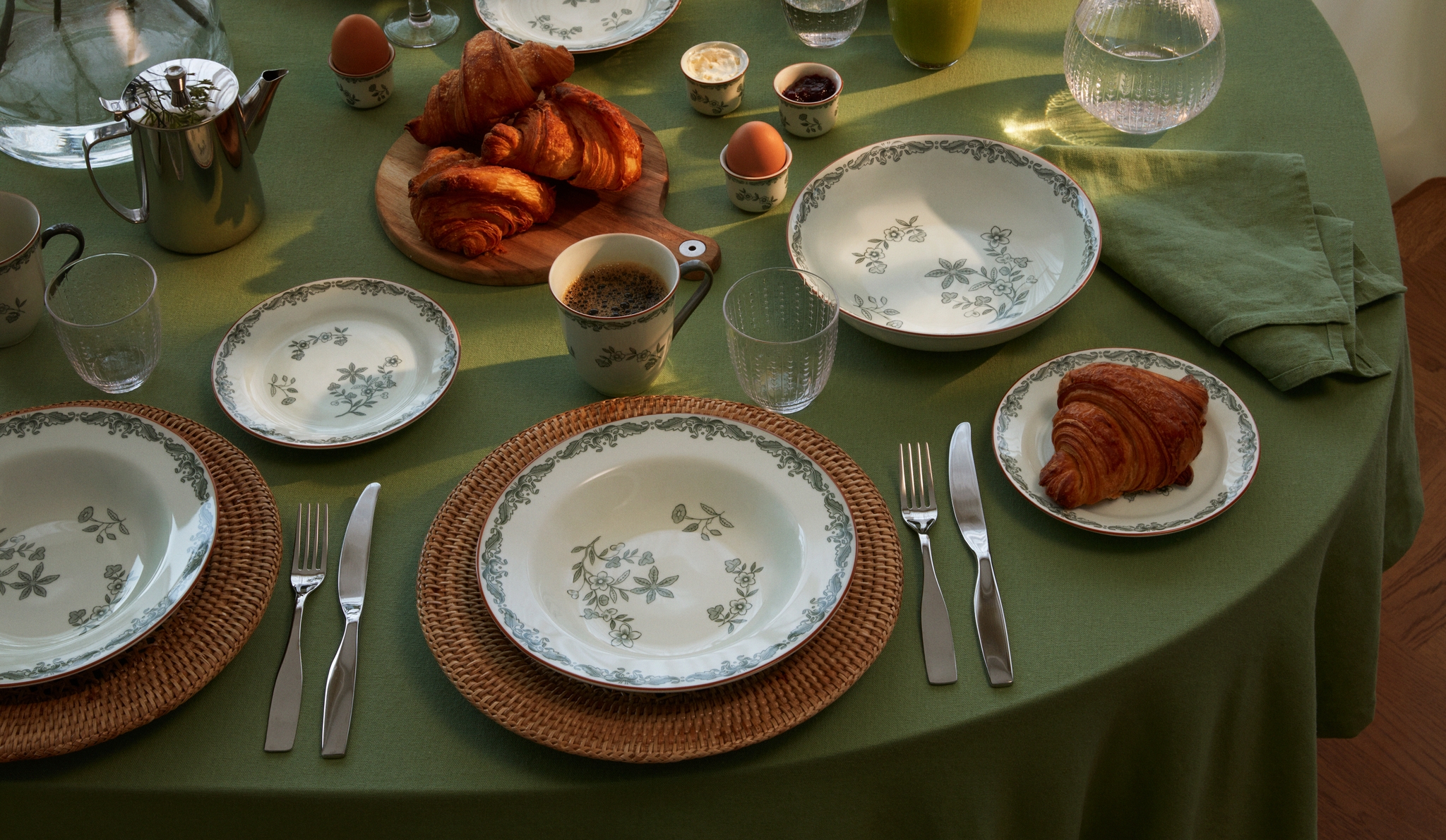 Frokostbord med grønn duk, blomstrete servise, croissanter, egg, kaffe og juice.