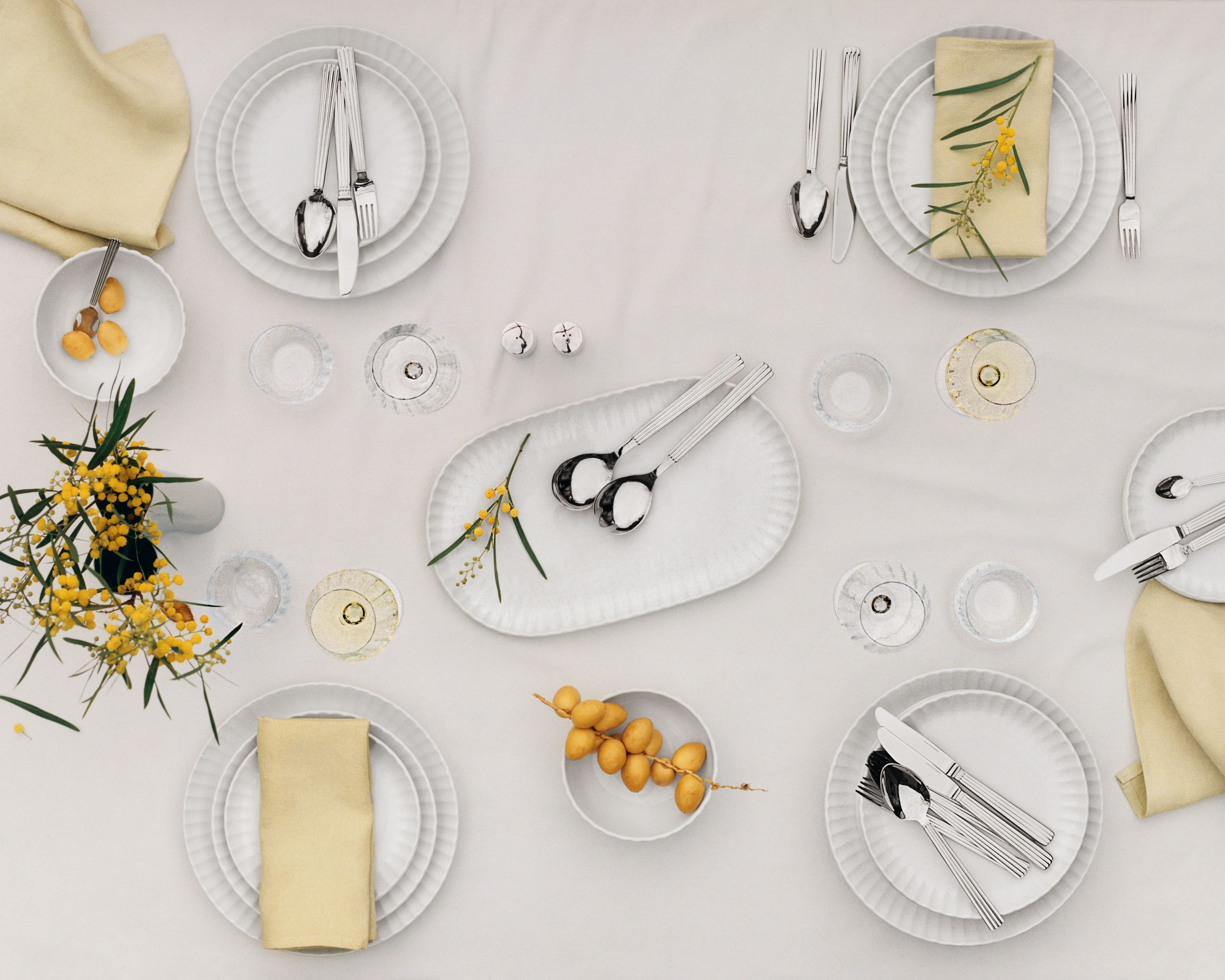  Ovanifrånvy av ett elegant matbord dukat med vita tallrikar, beige servetter, moderna bestick, vinglas och gula blommor.