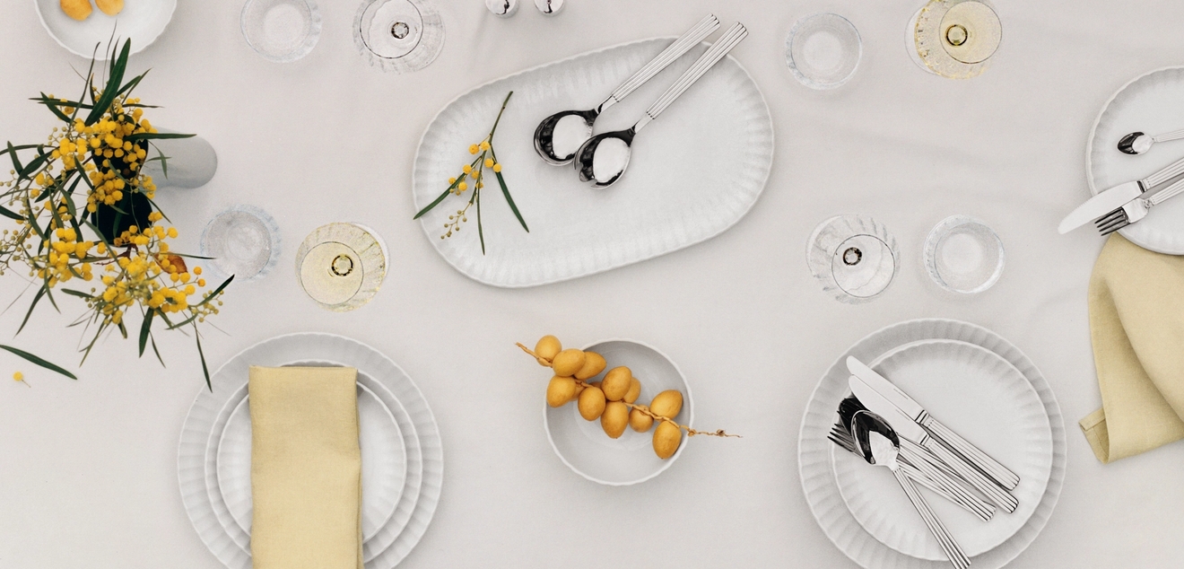 Ovanifrånvy av ett elegant matbord dukat med vita tallrikar, beige servetter, moderna bestick, vinglas och gula blommor.